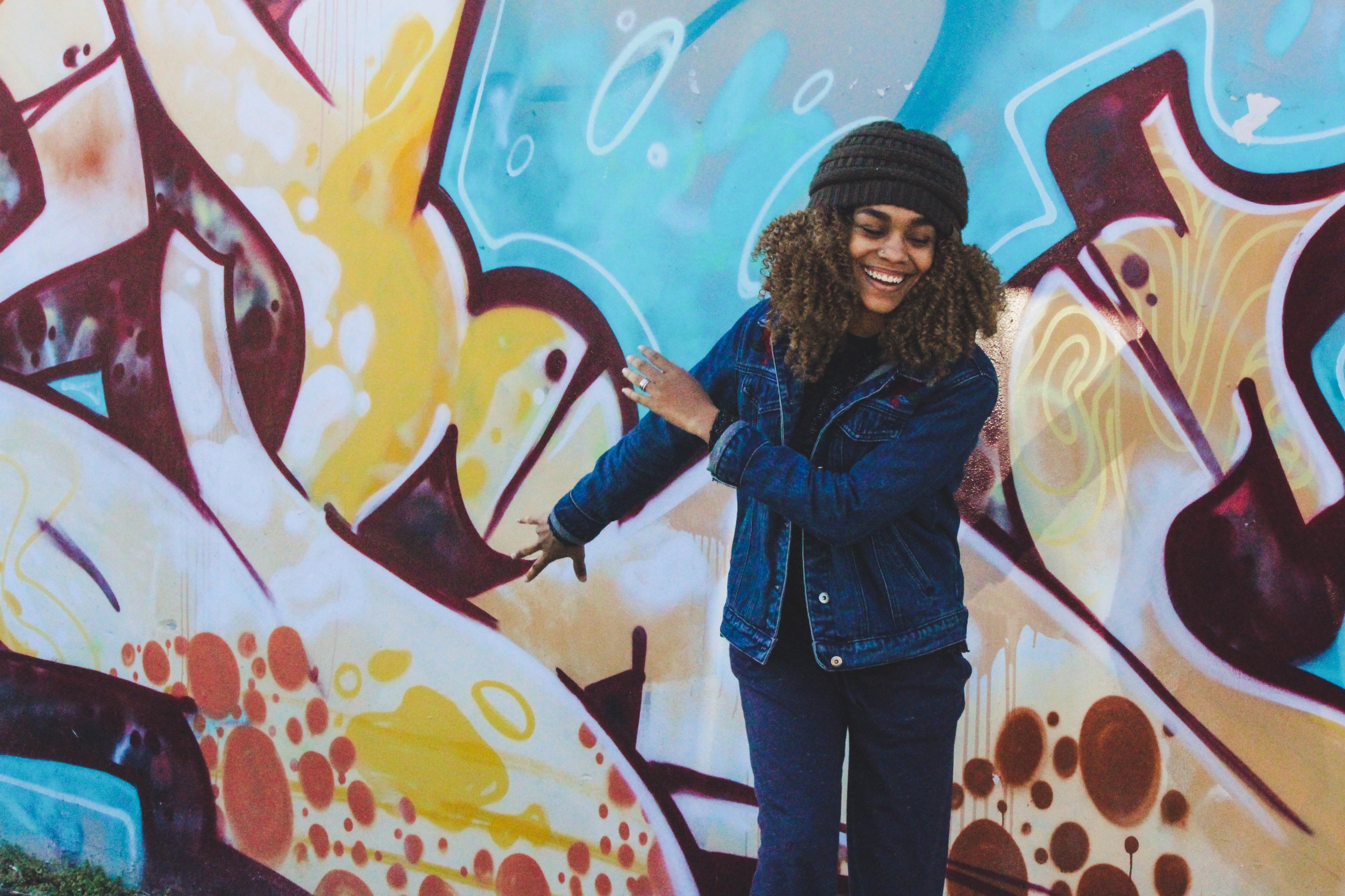 young woman wearing denim jacket and beanie smiling in front of bright graffiti wall, woman smiling in front of graffiti 4k, HD Wallpaper