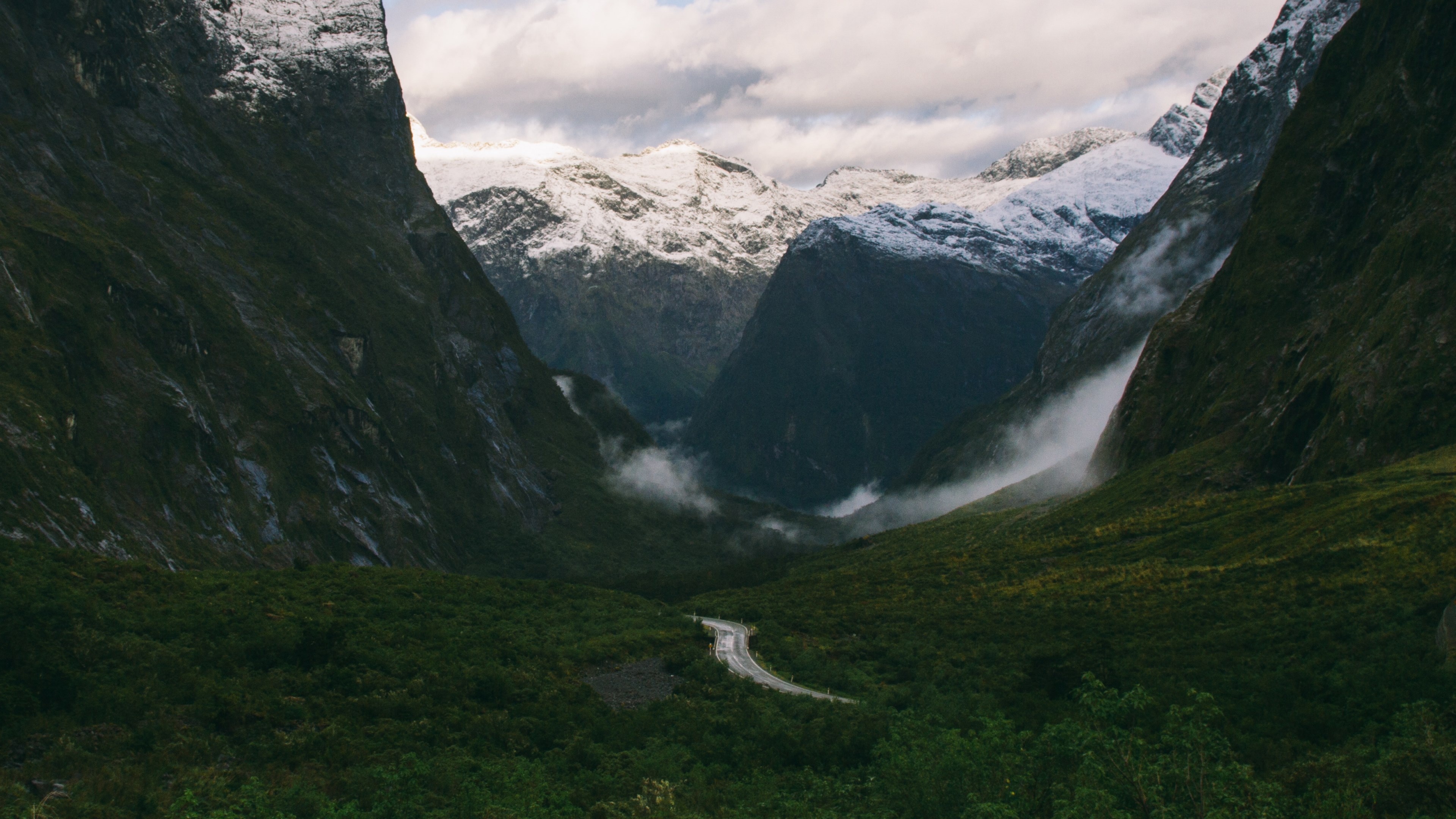 a narrow asphalt road running down a green mountain slope between snow capped ridges, road into the valleys 4k, HD Wallpaper
