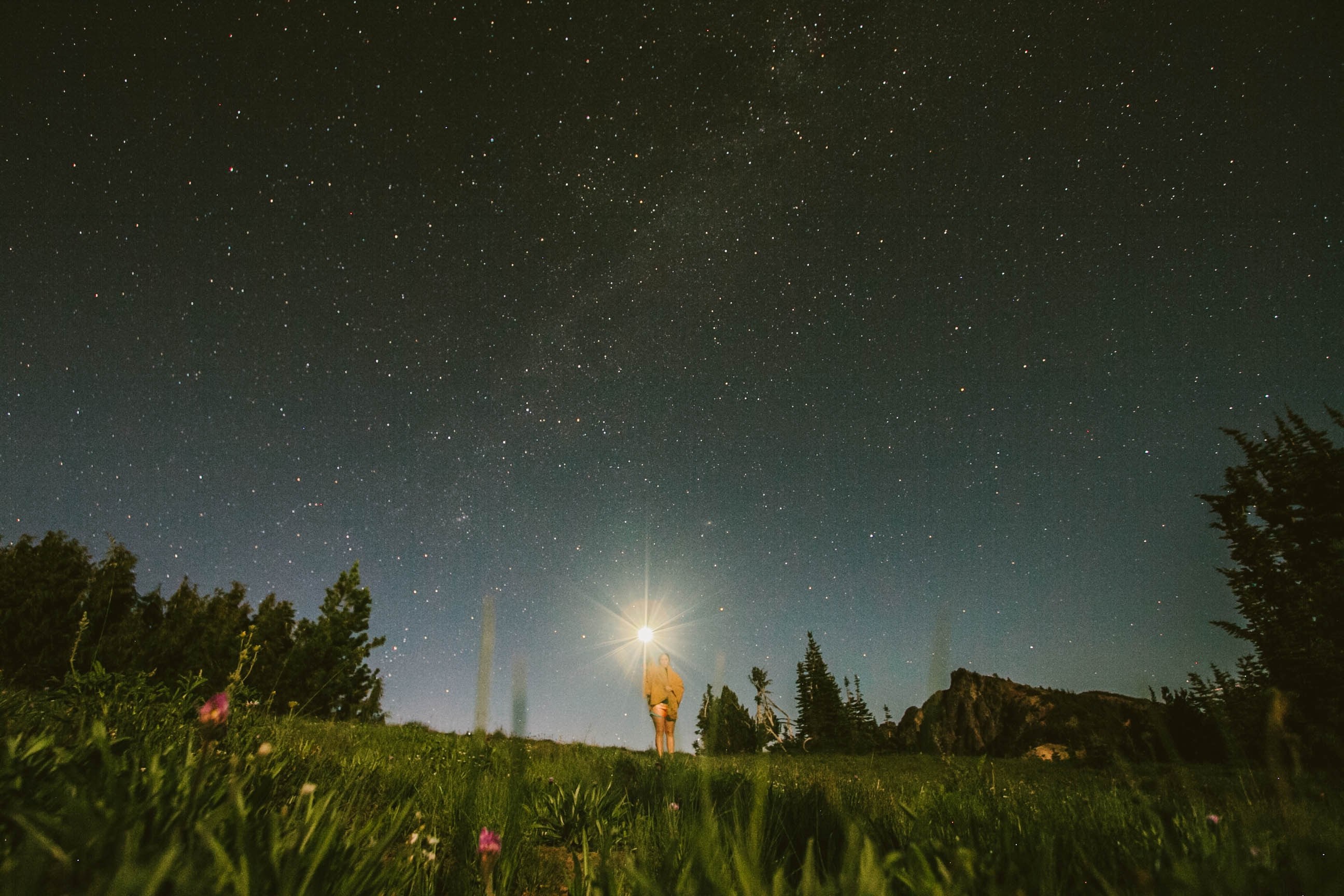 a starry night sky and a woman standing in the grass wearing a yellow blanket and holding a flashlight, lights in the dark 4k, HD Wallpaper