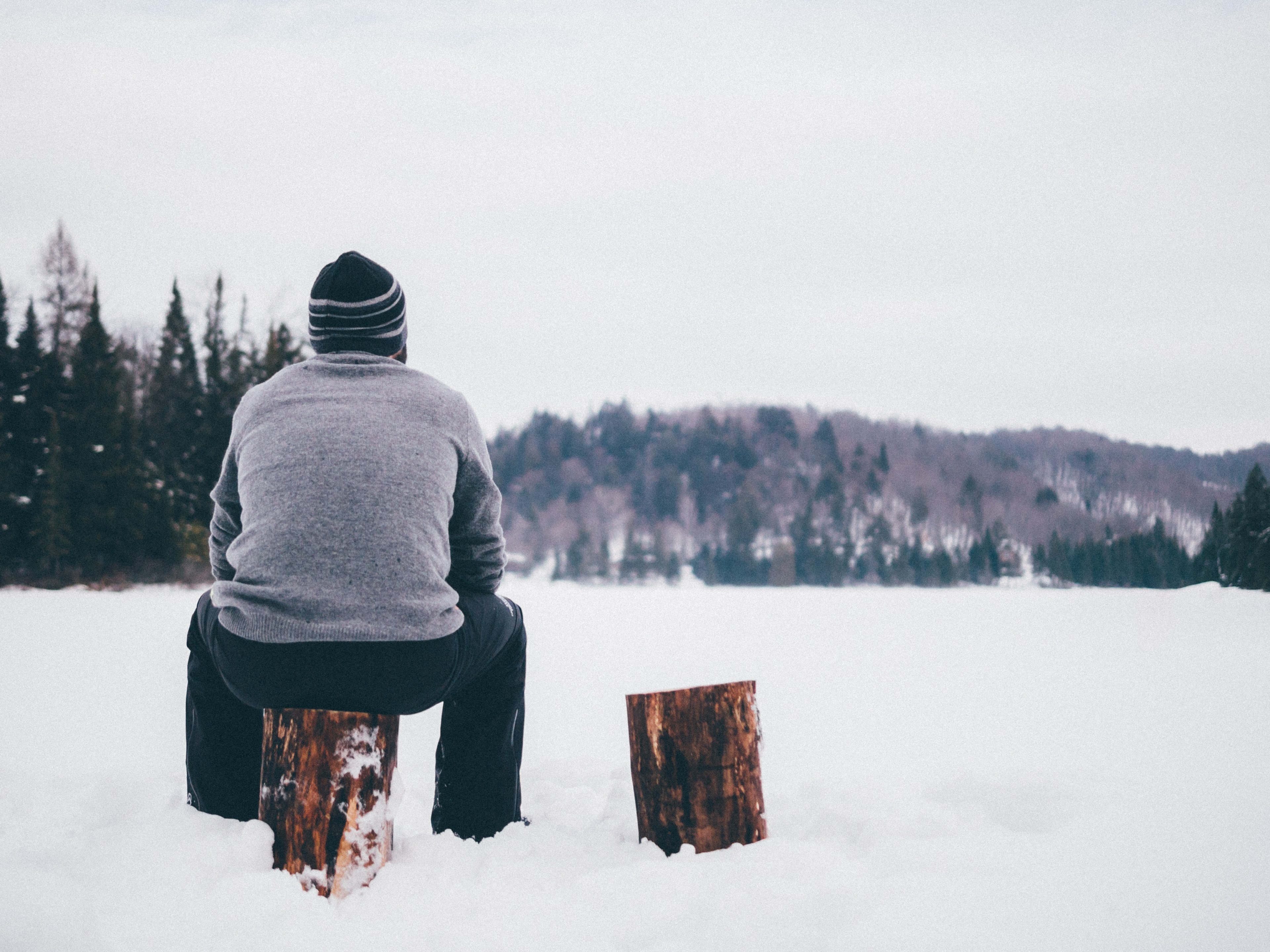a man wearing a knit cap and gray sweater looks out into a snowy forest while sitting on a log, man sits on log in snow 4k, HD Wallpaper