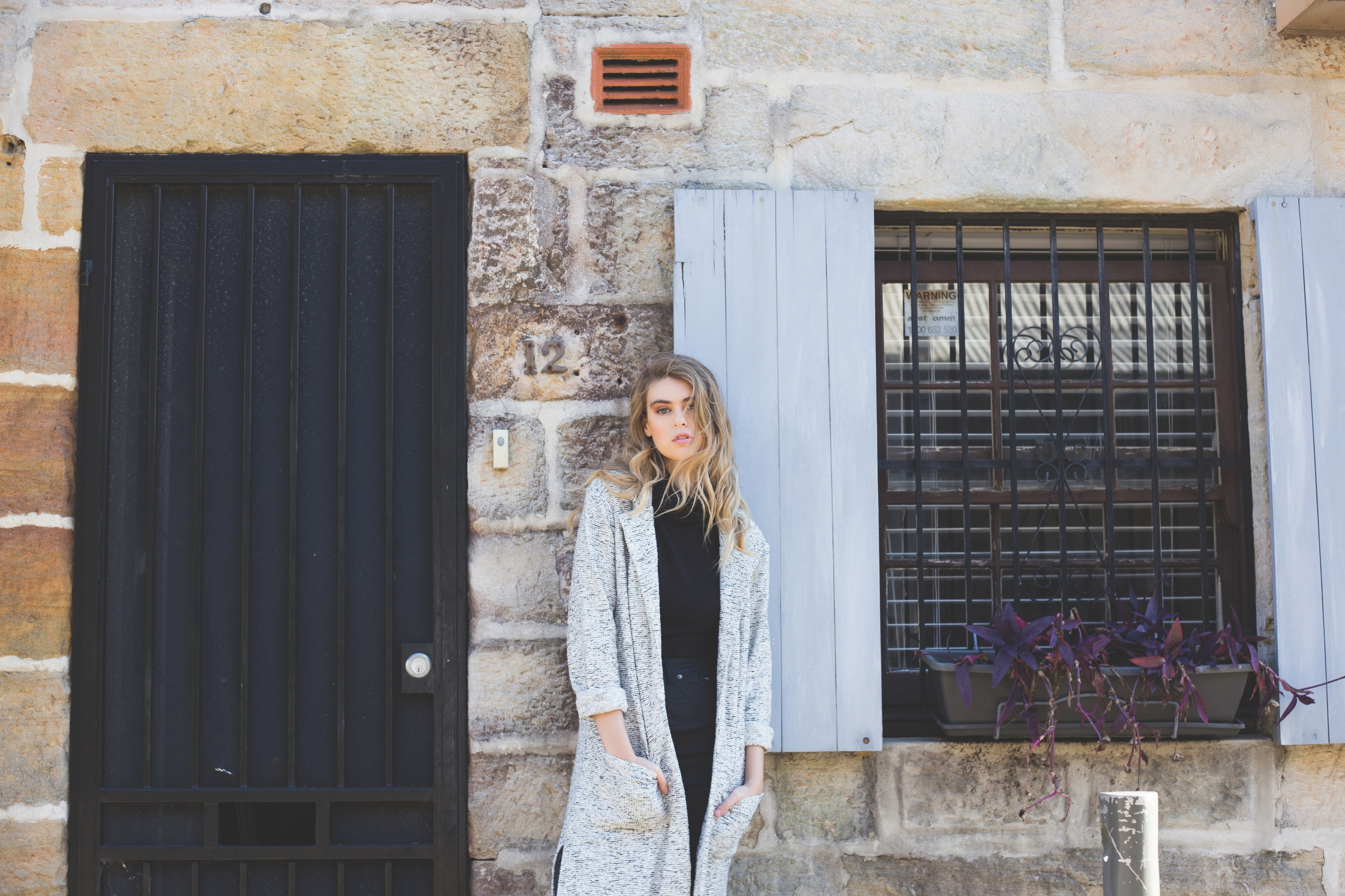 a woman in a long sweater stands in front of a stone front building with open shutters, woman and stone front building 4k, HD Wallpaper