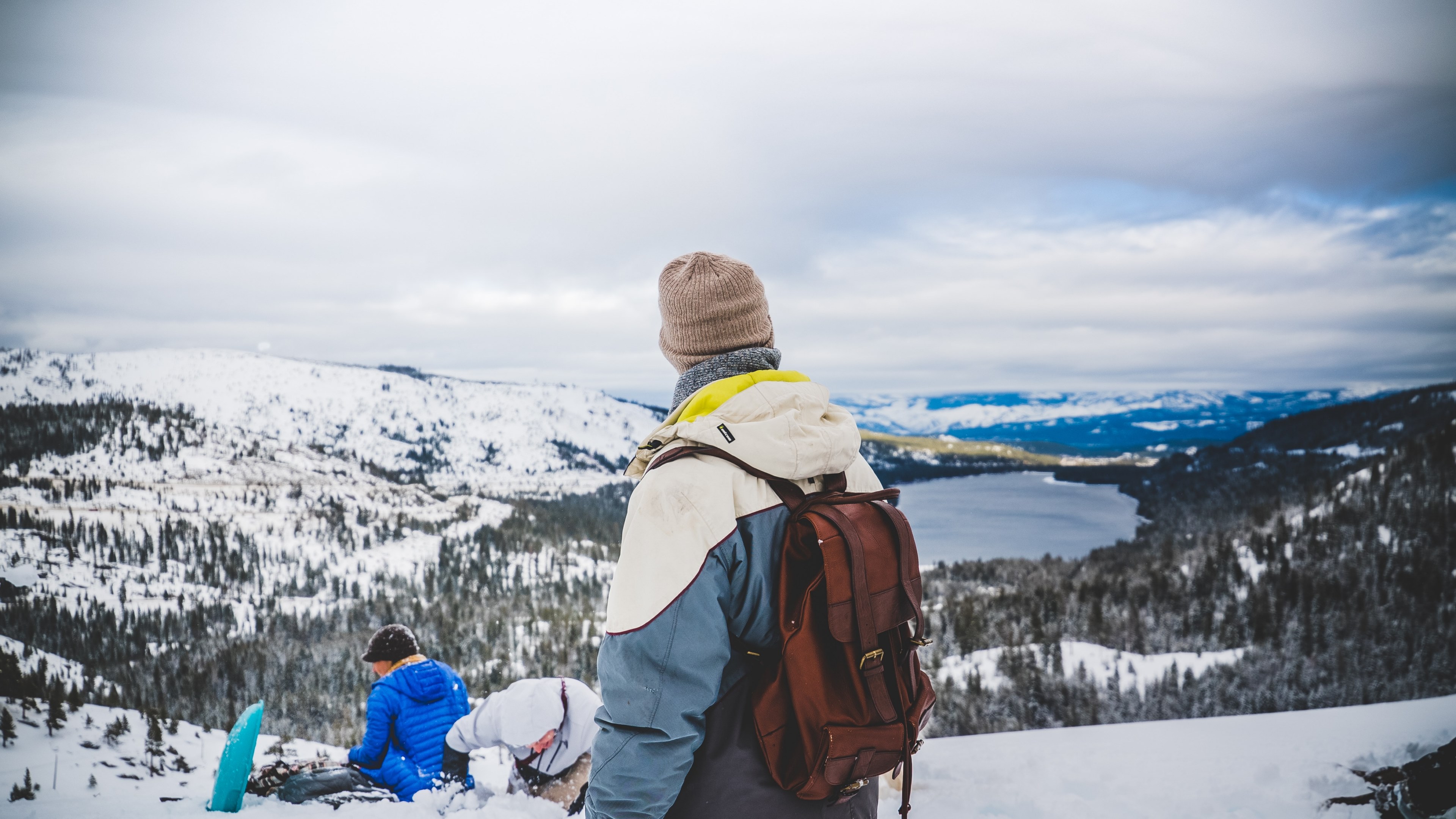 a backpacker in a beanie and a jacket looking at a snowy mountain valley with two people in the snow on his side, view on a mountain lake on a winters day 4k, HD Wallpaper