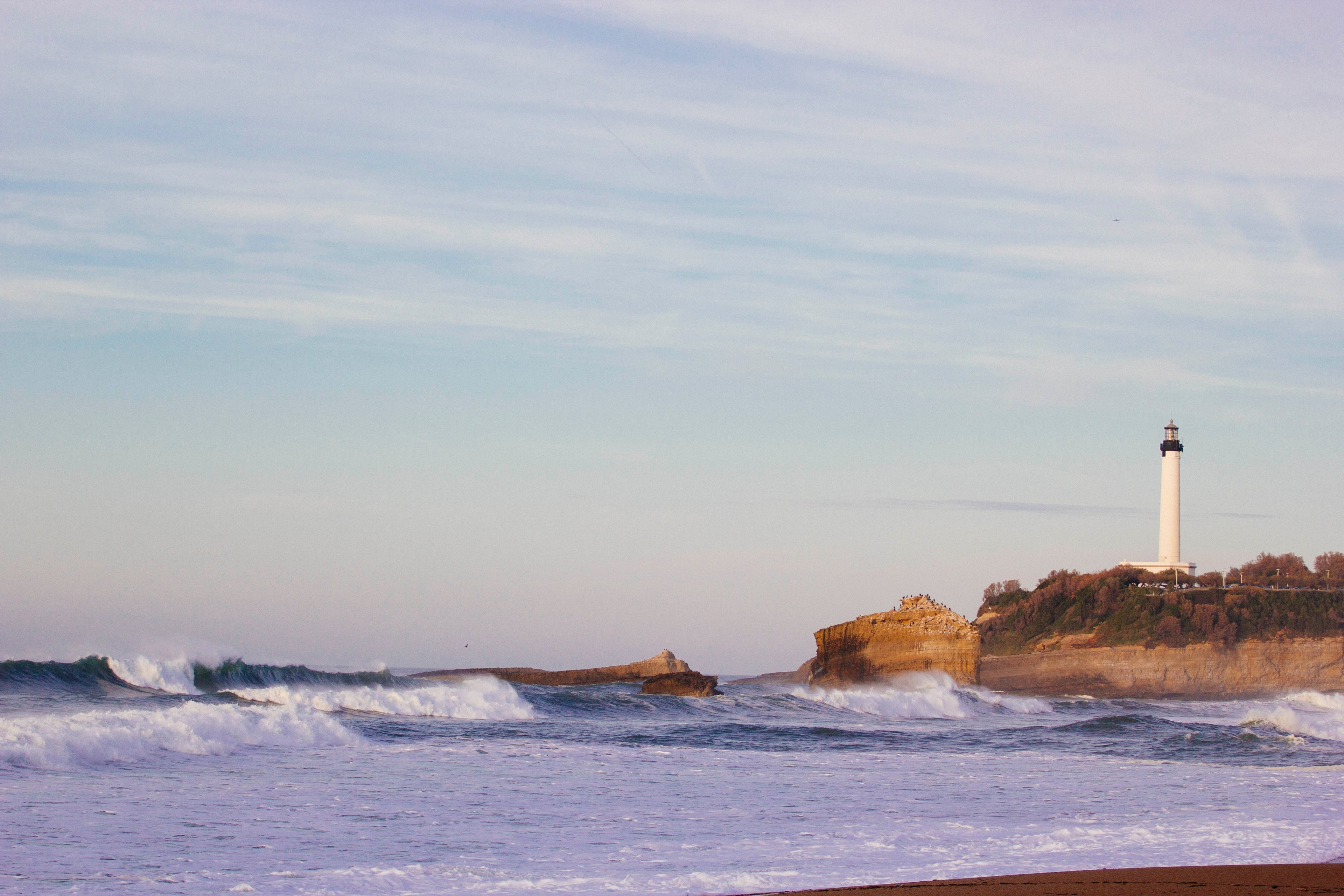 a coastal view of waves lapping the shoreline with a white lighthouse in the distance at biarritz, distant lighthouse 4k, HD Wallpaper