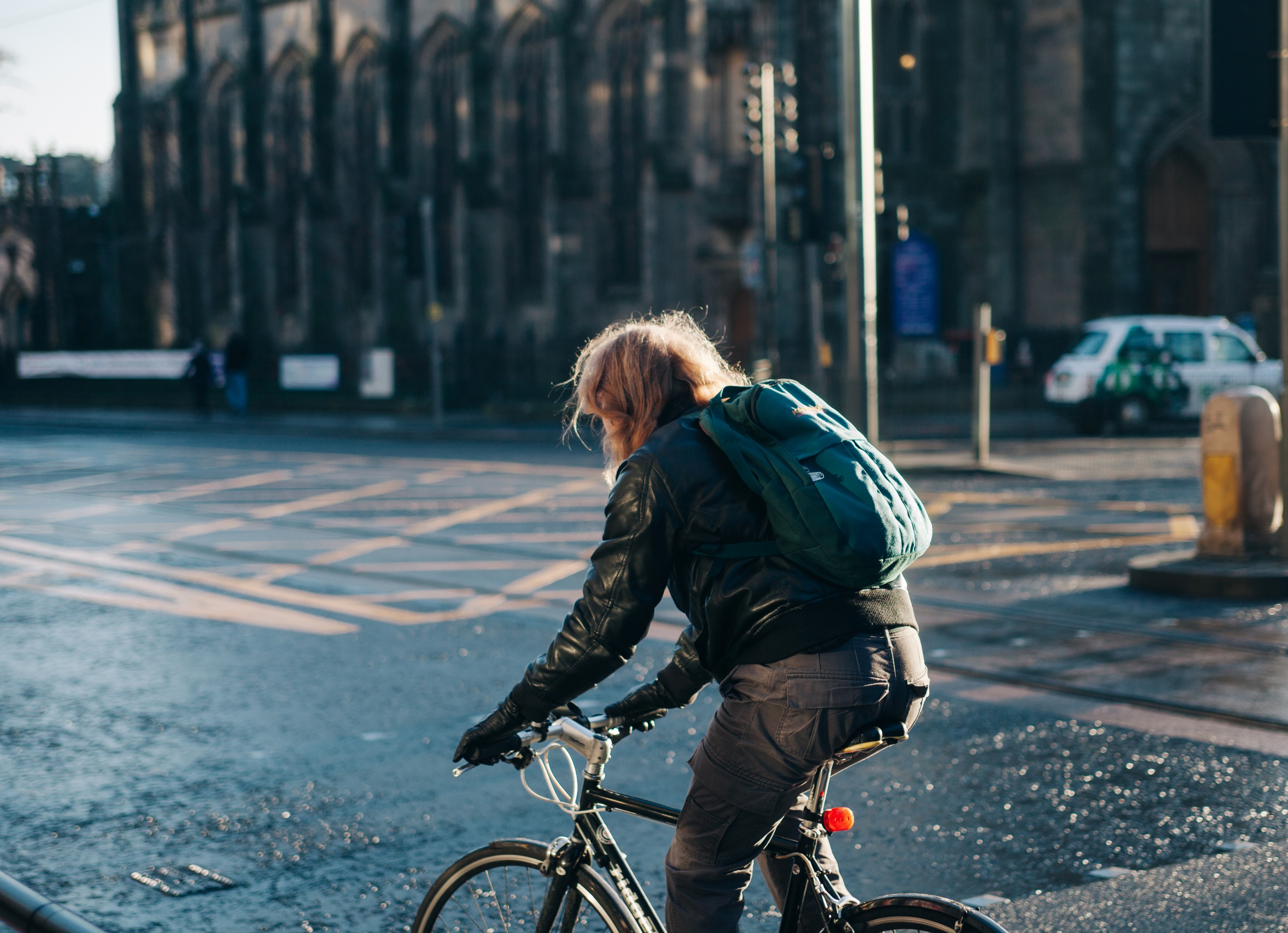 a person wearing a leather jacket and backpack riding a bicycle on a road in edinburgh, biking through the city 4k, HD Wallpaper