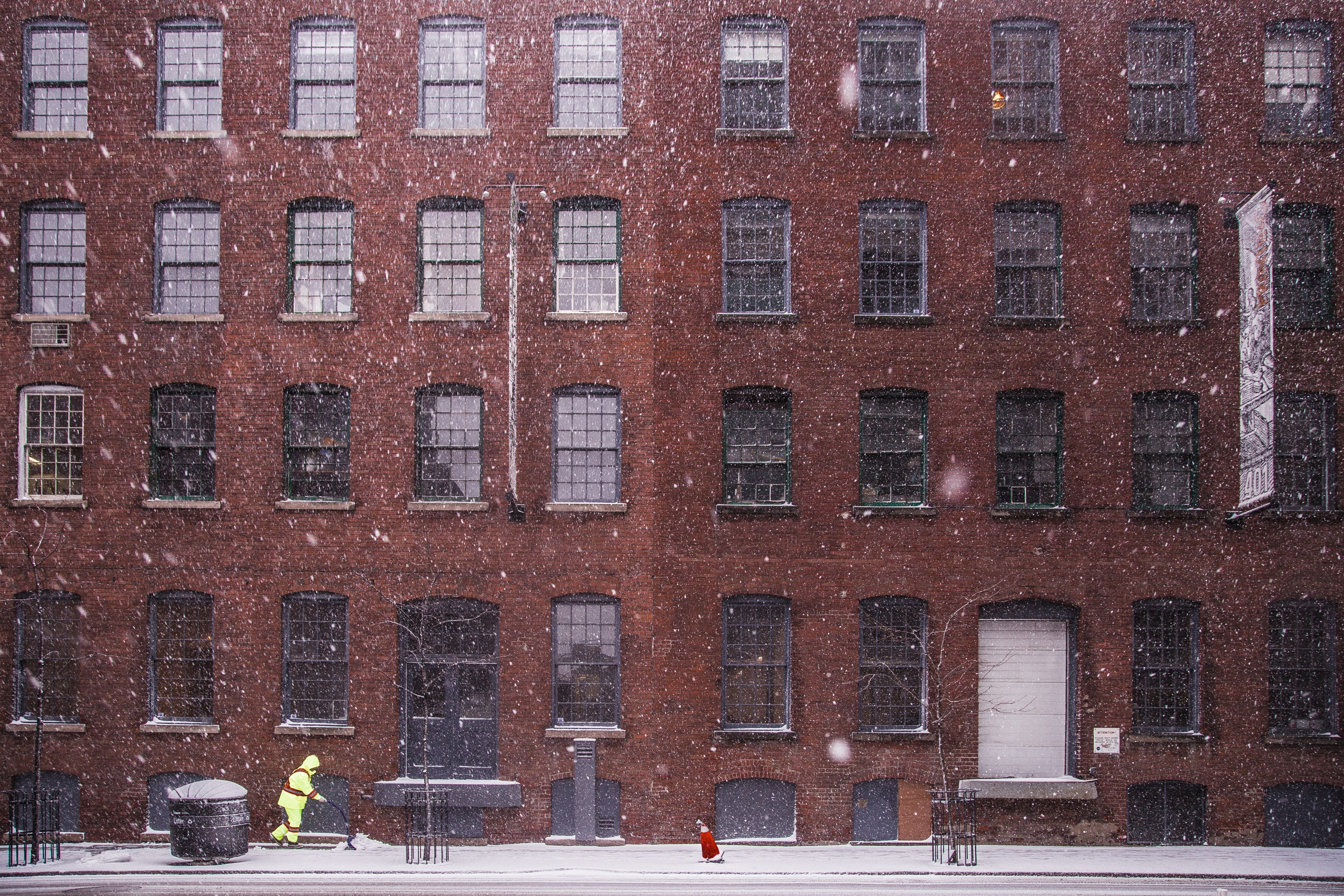 a person bundled in reflective yellow clothing shovels a sidewalk in front of a large red brick building during a snowfall, person shovels during snowfall 4k, HD Wallpaper