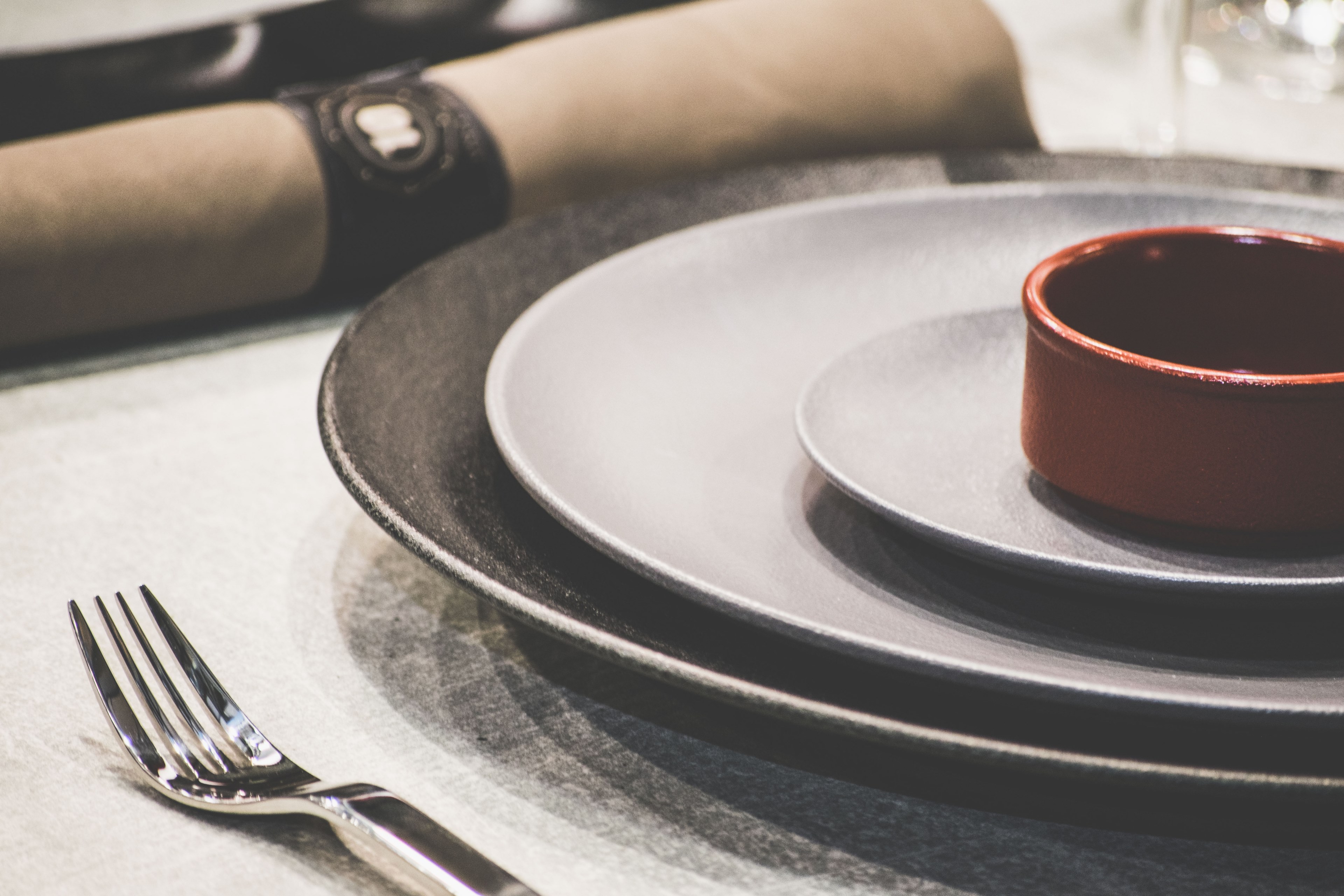 a red soup bowl on black and gray ceramic plates beside a shiny silver fork and a rolled up napkin, dinnerware setup 4k, HD Wallpaper