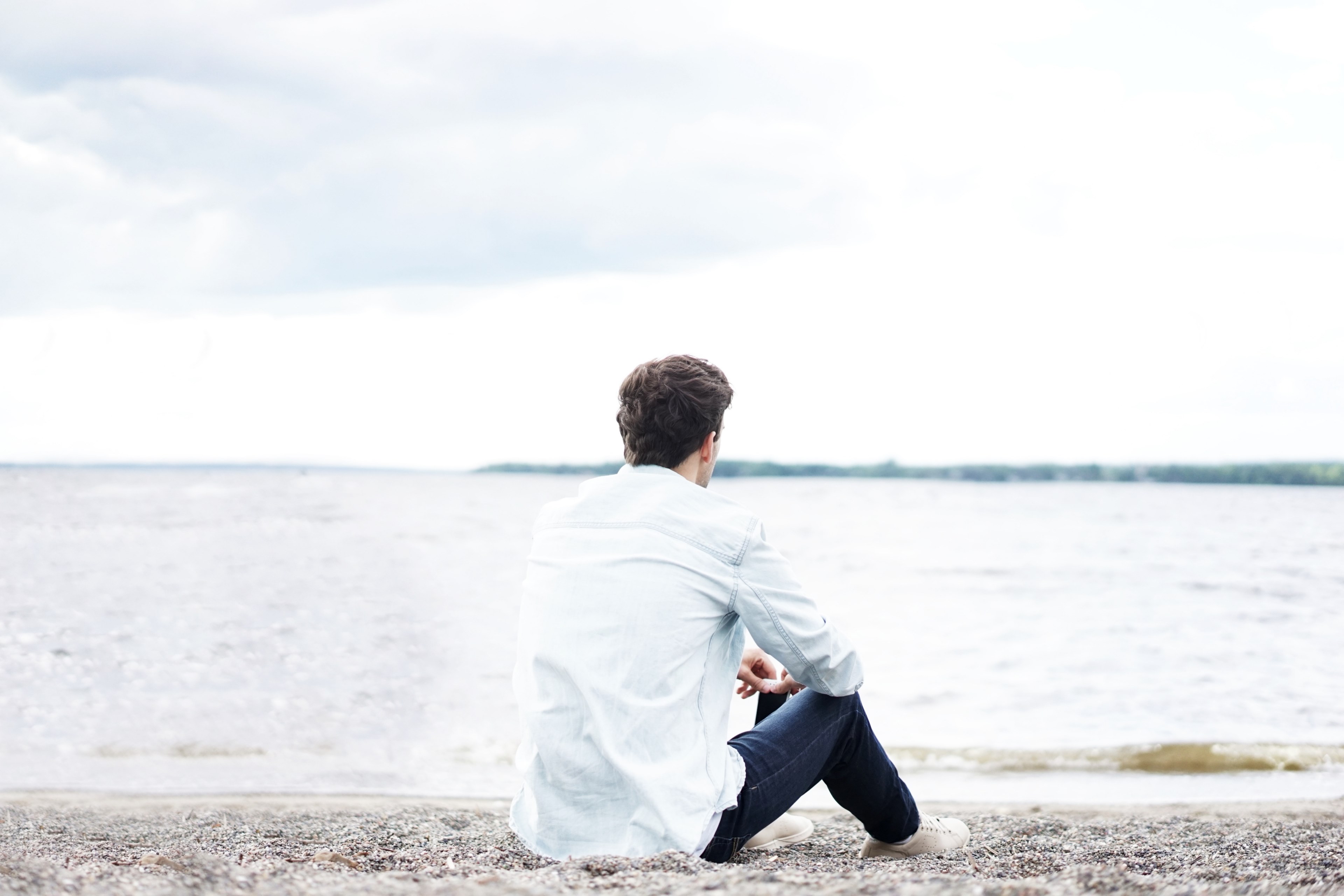 a man in a white shirt sitting on a pebble beach and looking of into the sea, contemplation on a beach 4k, HD Wallpaper