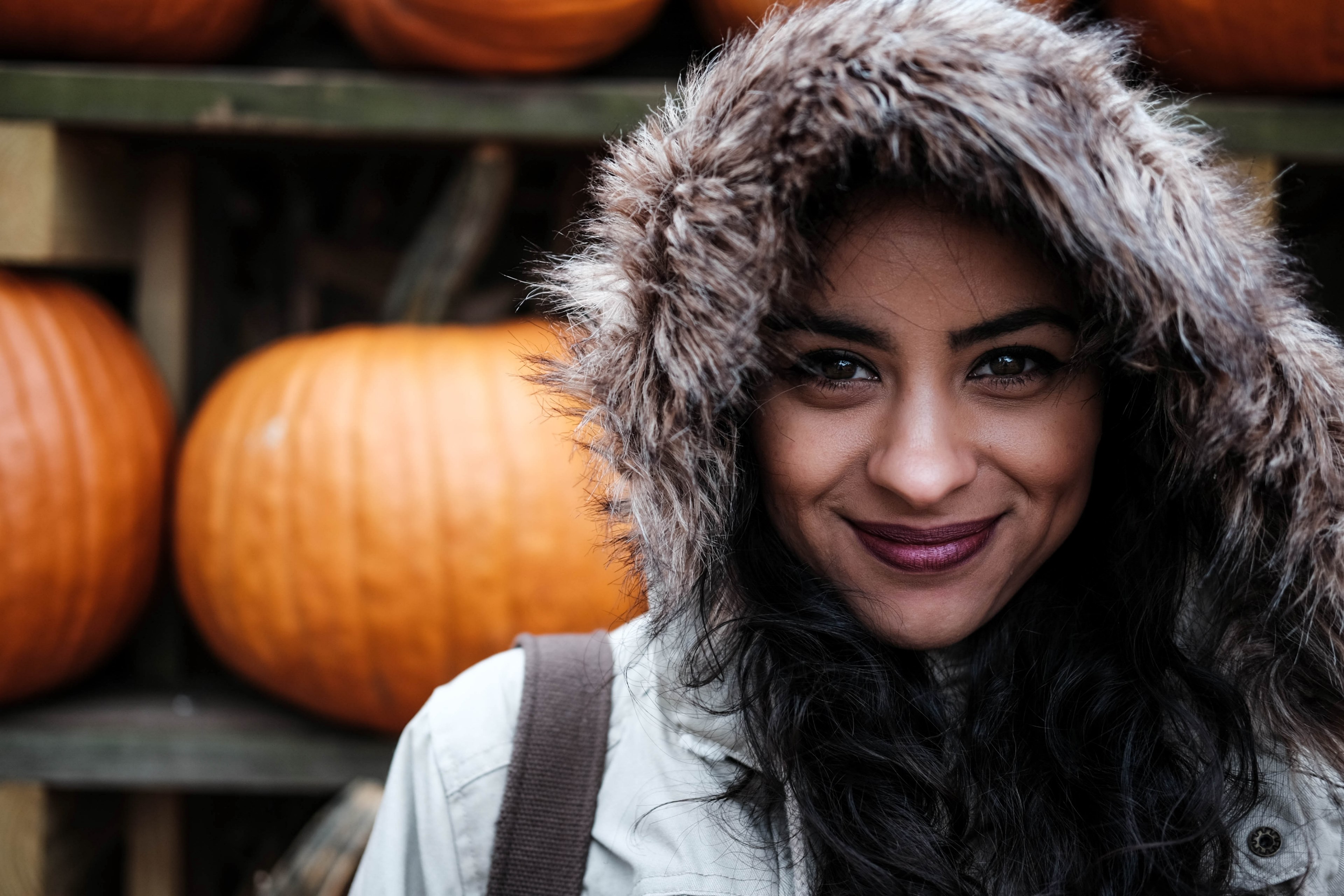 a woman wearing a fur hood and maroon lipstick smiles in front of shelves of pumpkins, in front of pumpkin shelves 4k, HD Wallpaper