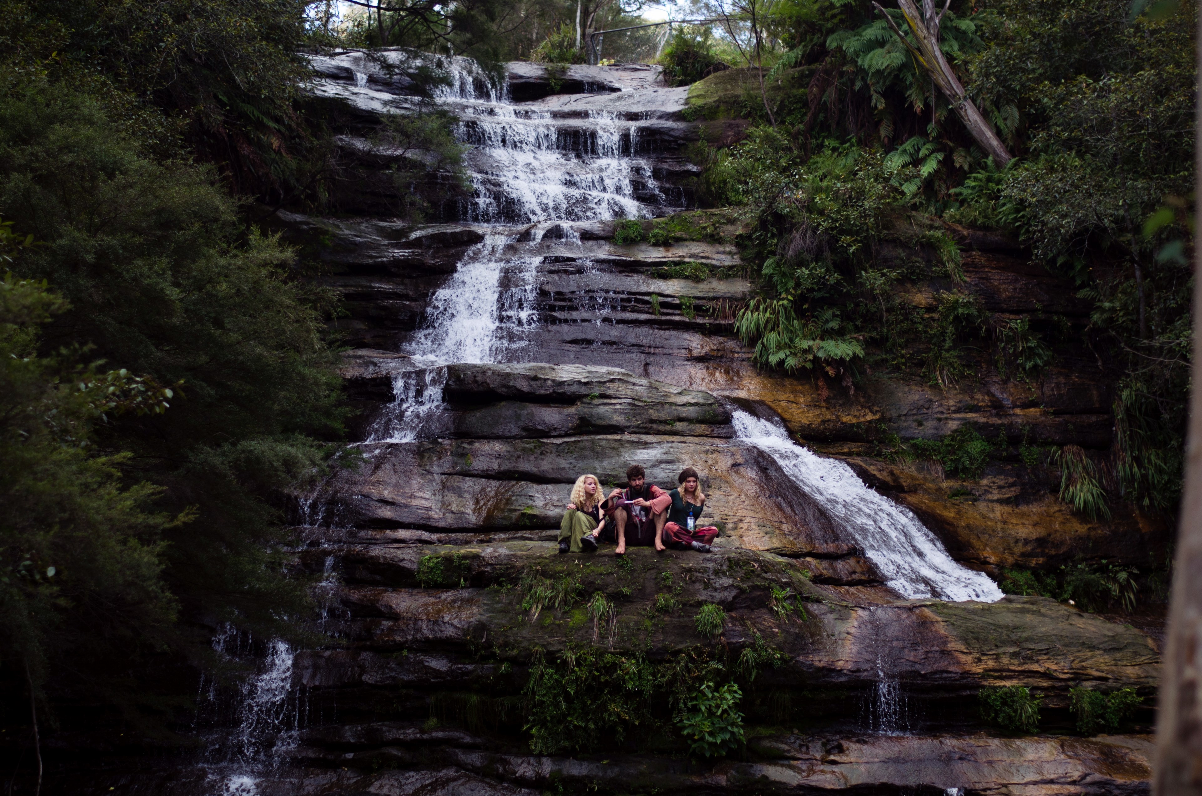 three young people sitting on a rocky ledge with a waterfall streaming down on their side, sitting on a waterfall ledge 4k, HD Wallpaper