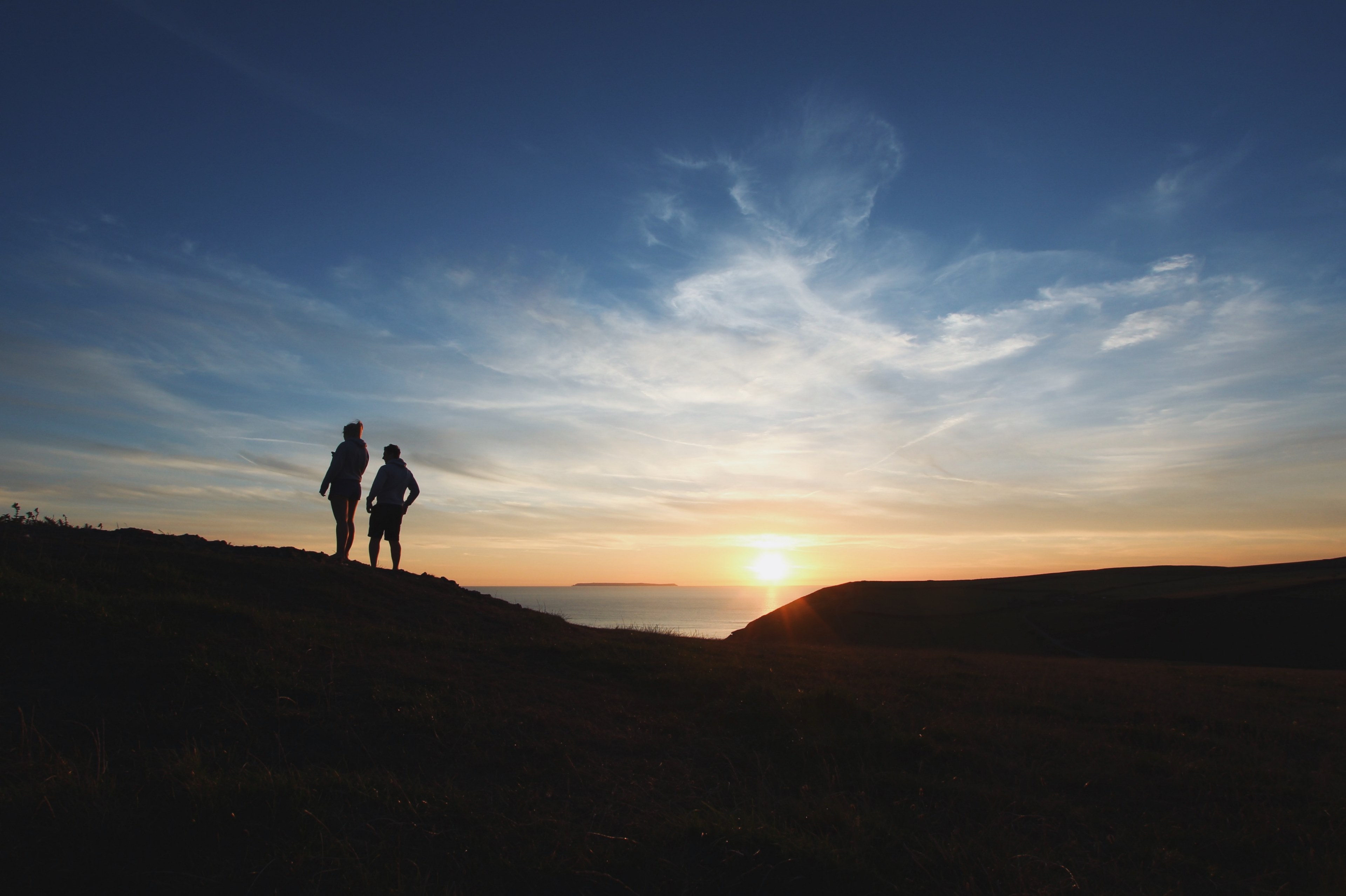 silhouette couple standing on a hill at croyde bay facing away from a distant sea view at sunrise or sunset causing a yellow sky turning blue covered in light and airy cloud, walking along 4k, HD Wallpaper