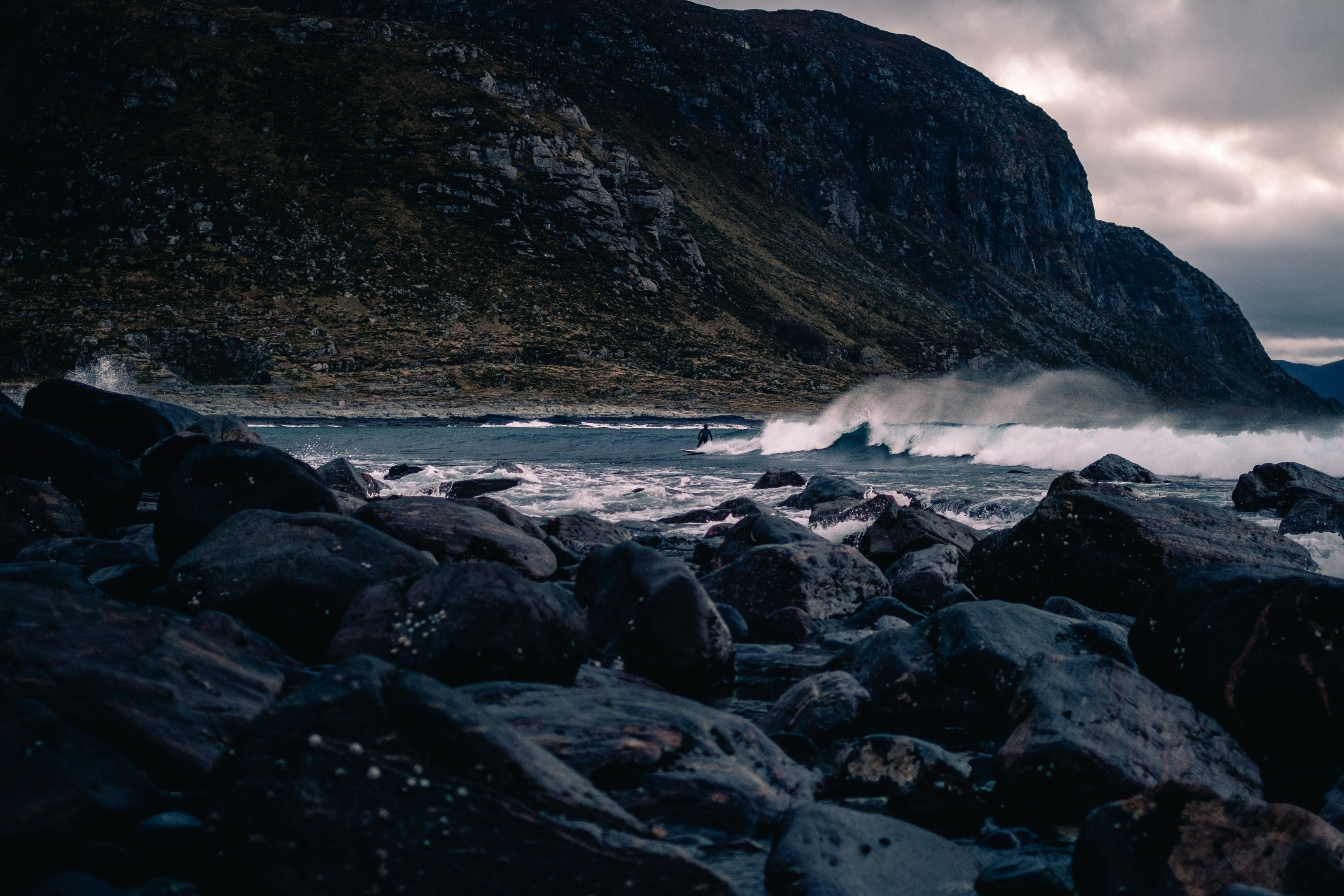a distant surfer riding the waves in front of a coastal cliff seen from the rocks on the shore at alnes, surfer and the rocks 4k, HD Wallpaper