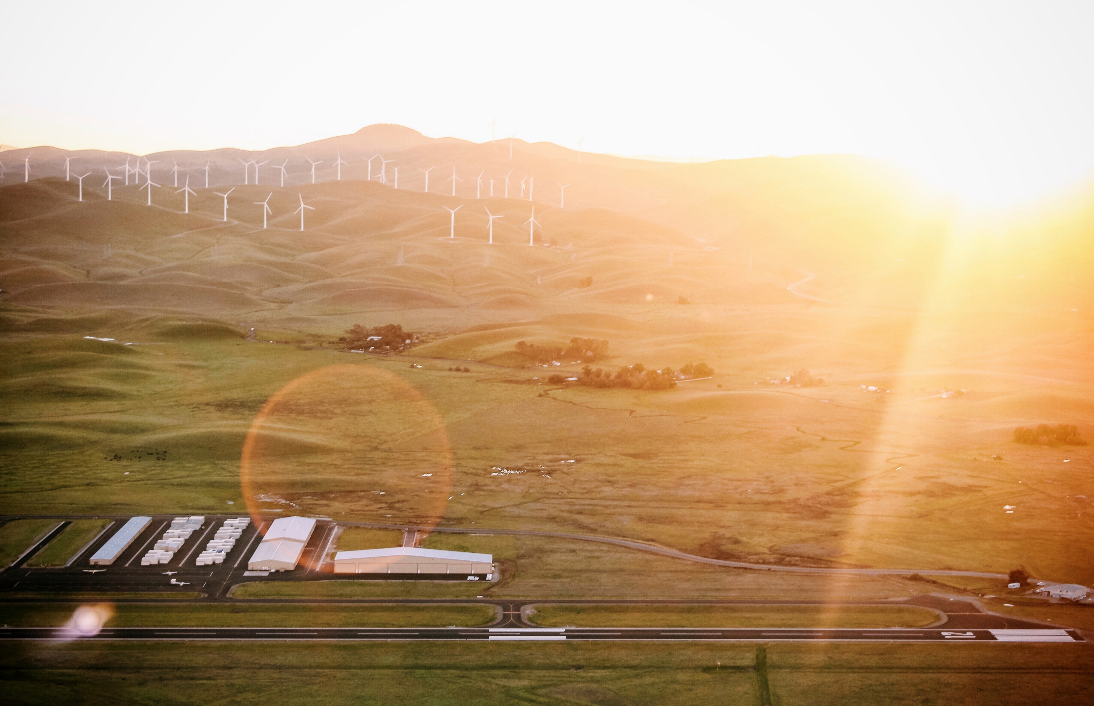 a drone view of byron airport the runway and hangers as well as the surrounding countryside including grassland hills and a wind farm with sun white glare of sunrise or sunset in the distanc 4k, HD Wallpaper