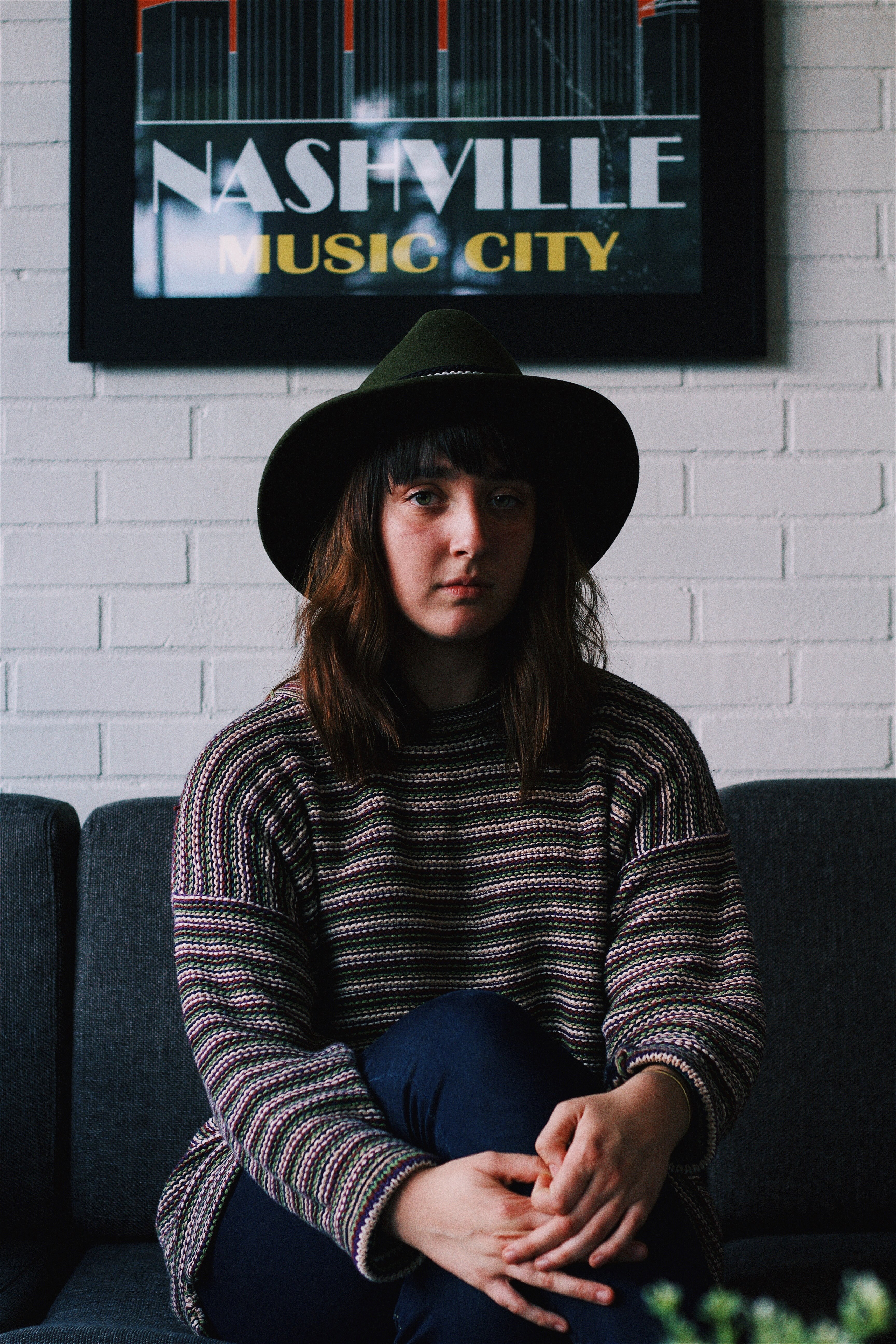 a young woman in a hat sitting on a sofa under a white wall with a nashville music city poster, woman under nashville music city poster 4k, Phone HD Wallpaper