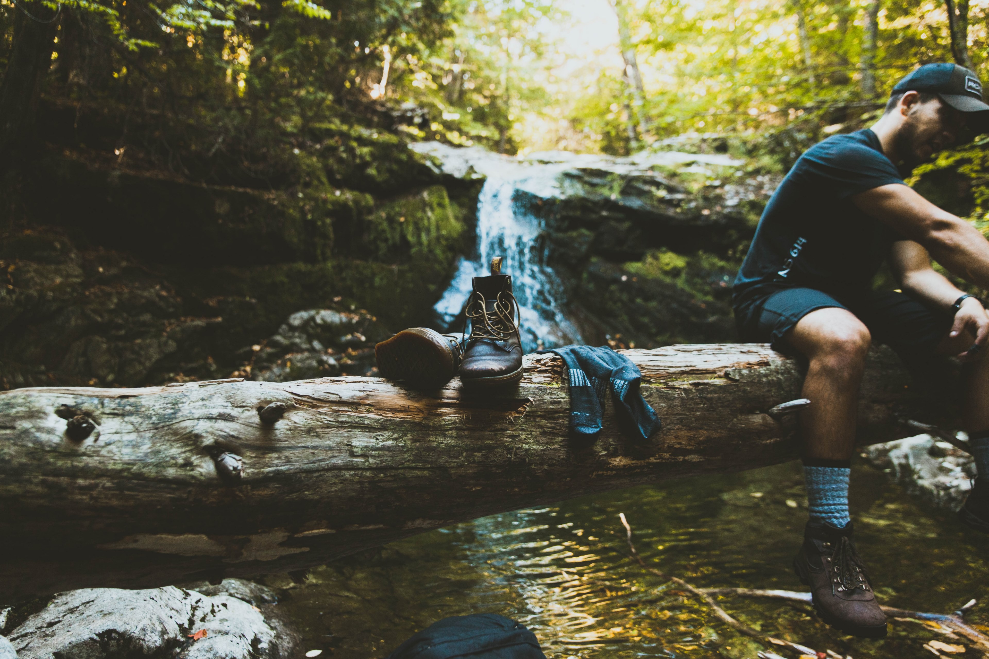 a hiker sitting on a fallen tree stump next to a pair of hiking boots in front of a waterfall, hiker resting on a log 4k, HD Wallpaper