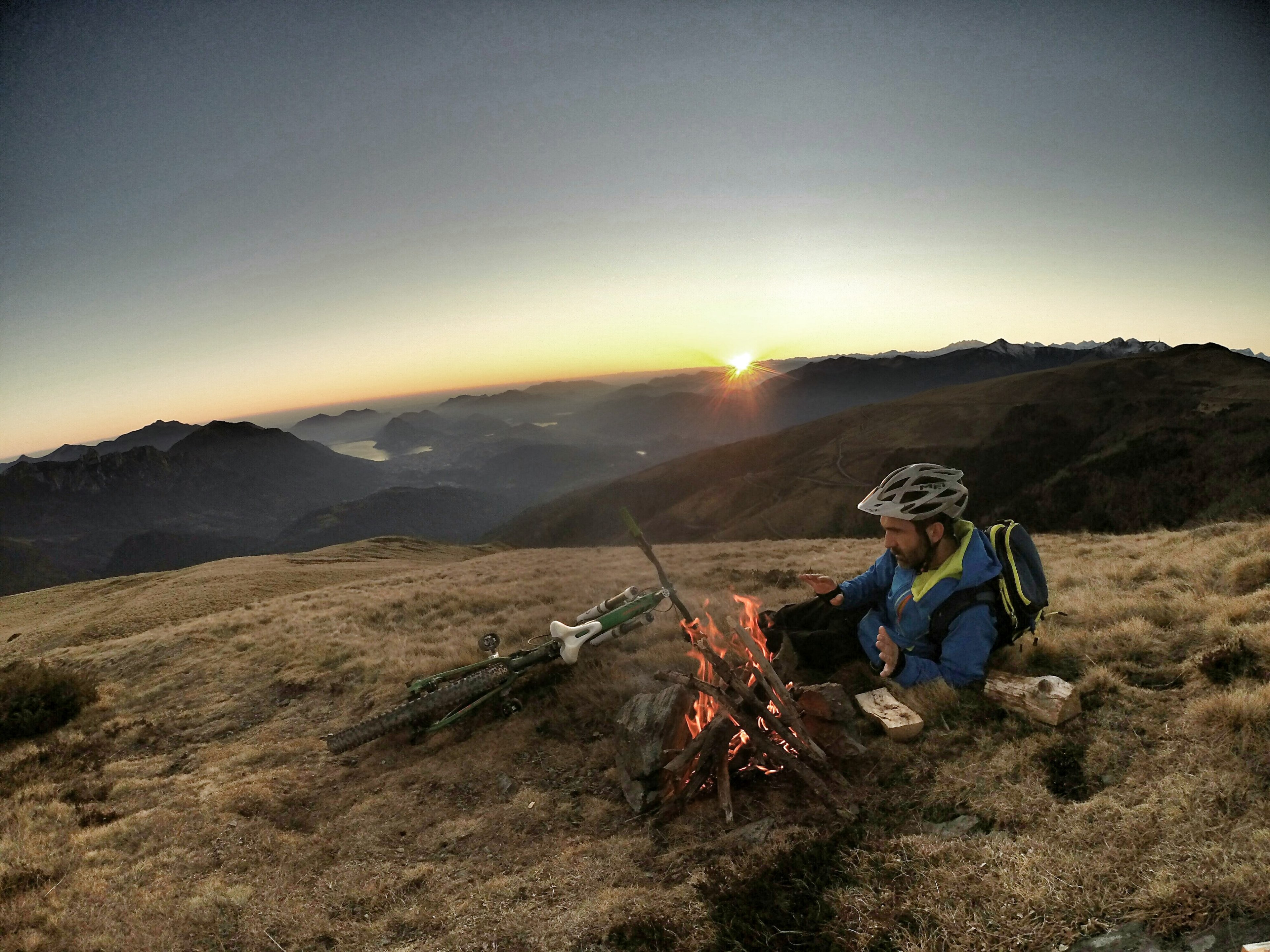 a man with a jacket and bike helmet on sitting by a fire and his bicycle in on a hill, cycling across the mountainside 4k, HD Wallpaper