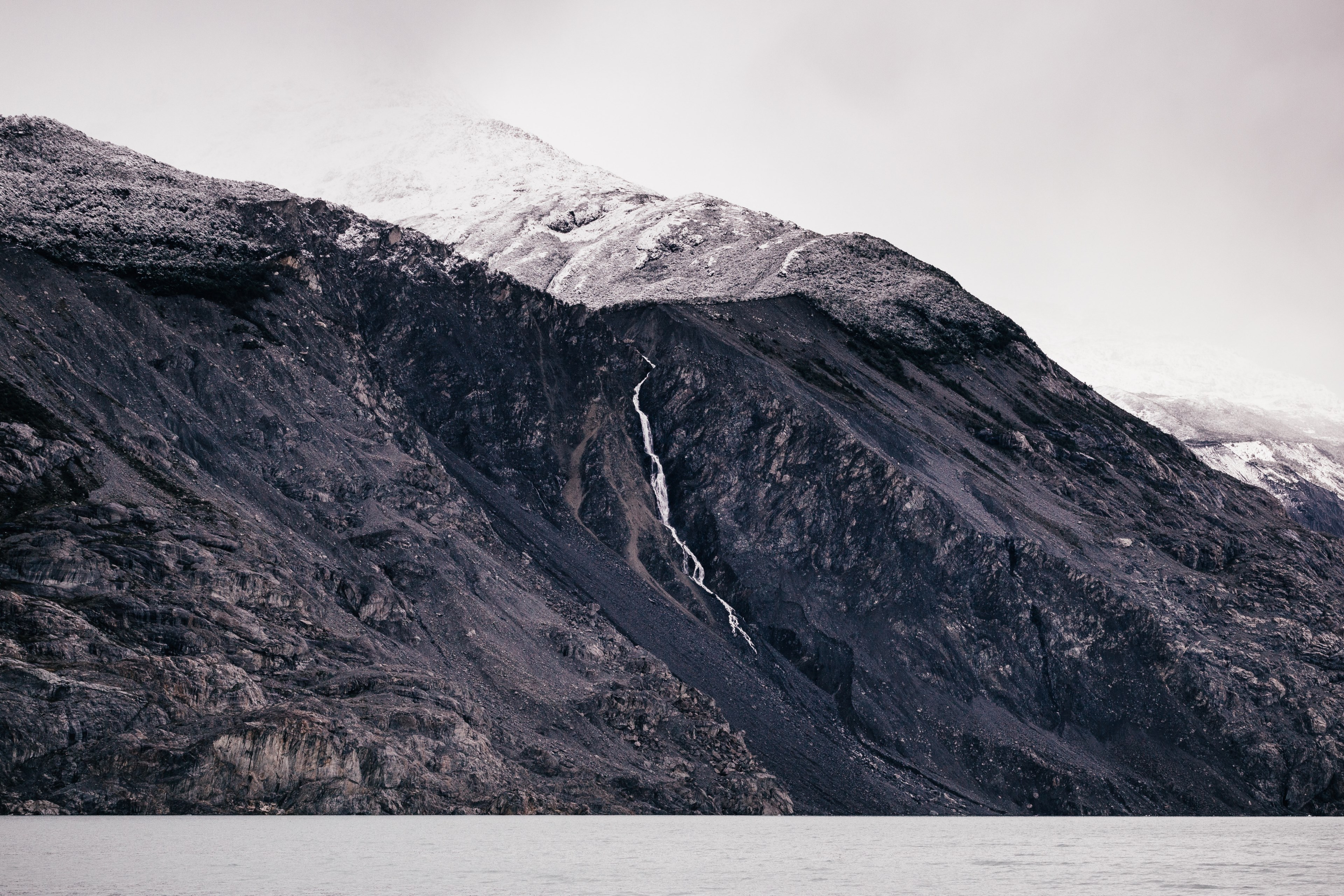 large rocky mountains with a snowy peak and a river running down an edge on a cloudy day, rocky mountain and river 4k, HD Wallpaper