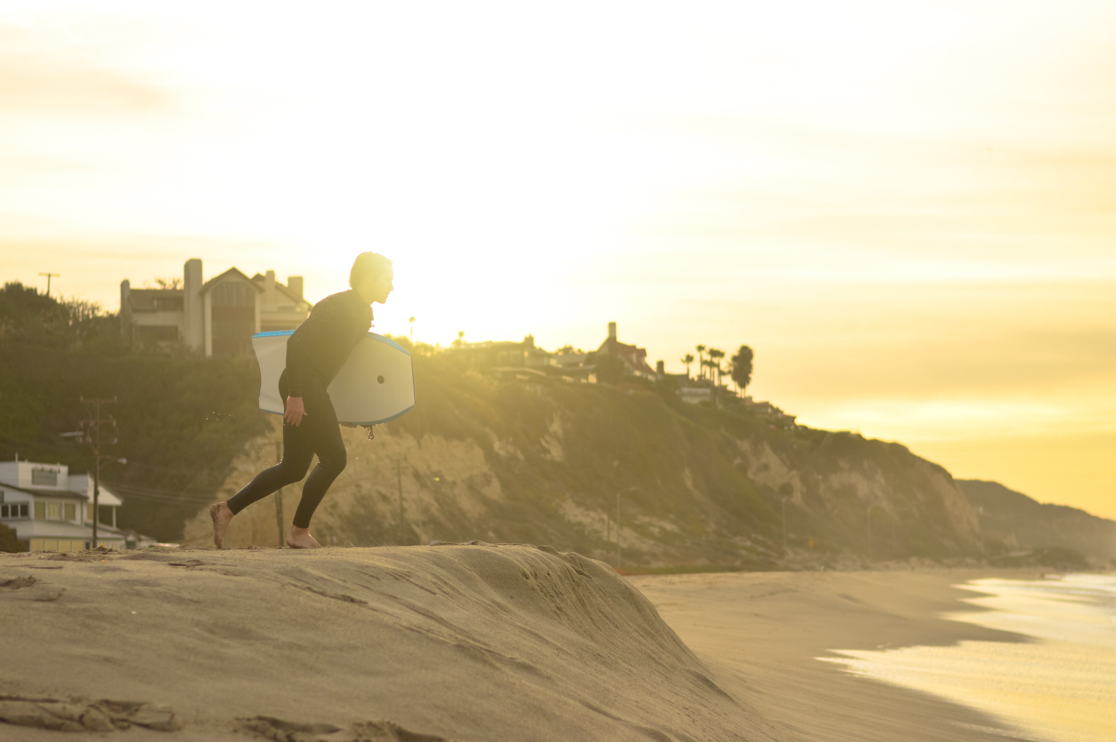 a surfer in a wetsuit with his surfboard under his arm running across the sand towards the sea at sunset, surfer in malibu 4k, HD Wallpaper