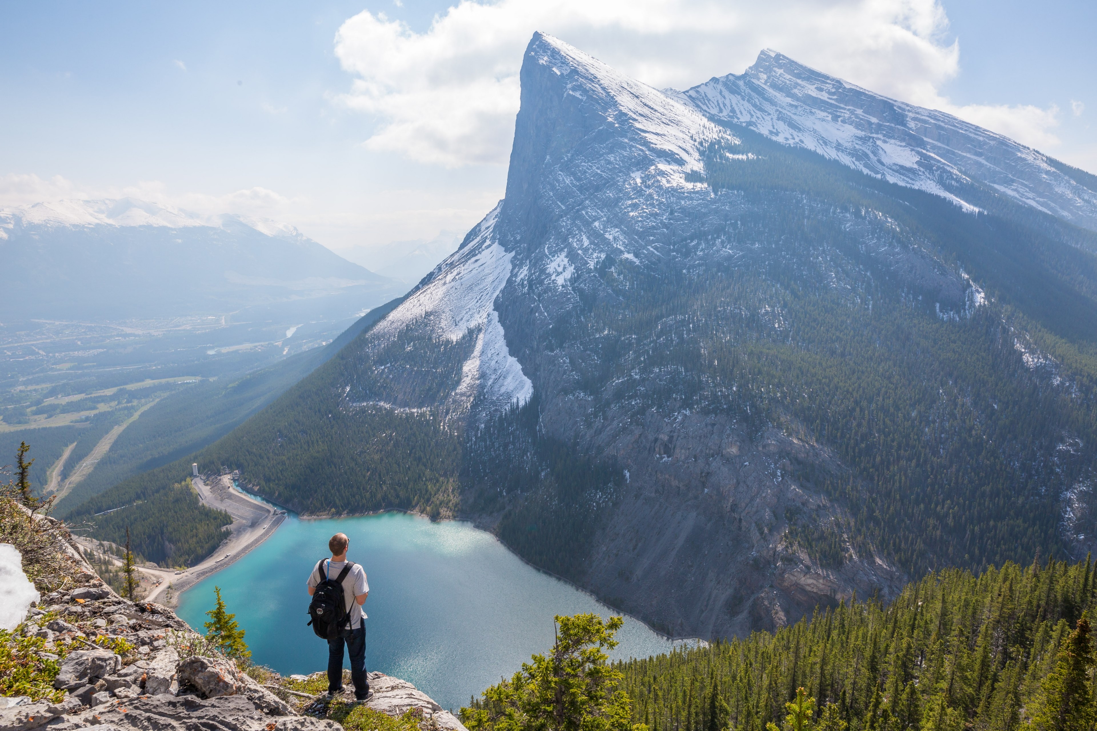 a man wearing a backpack and jeans looking out at the lake and mountains at east end of rundle eeor trail, backpacker on a high ledge 4k, HD Wallpaper