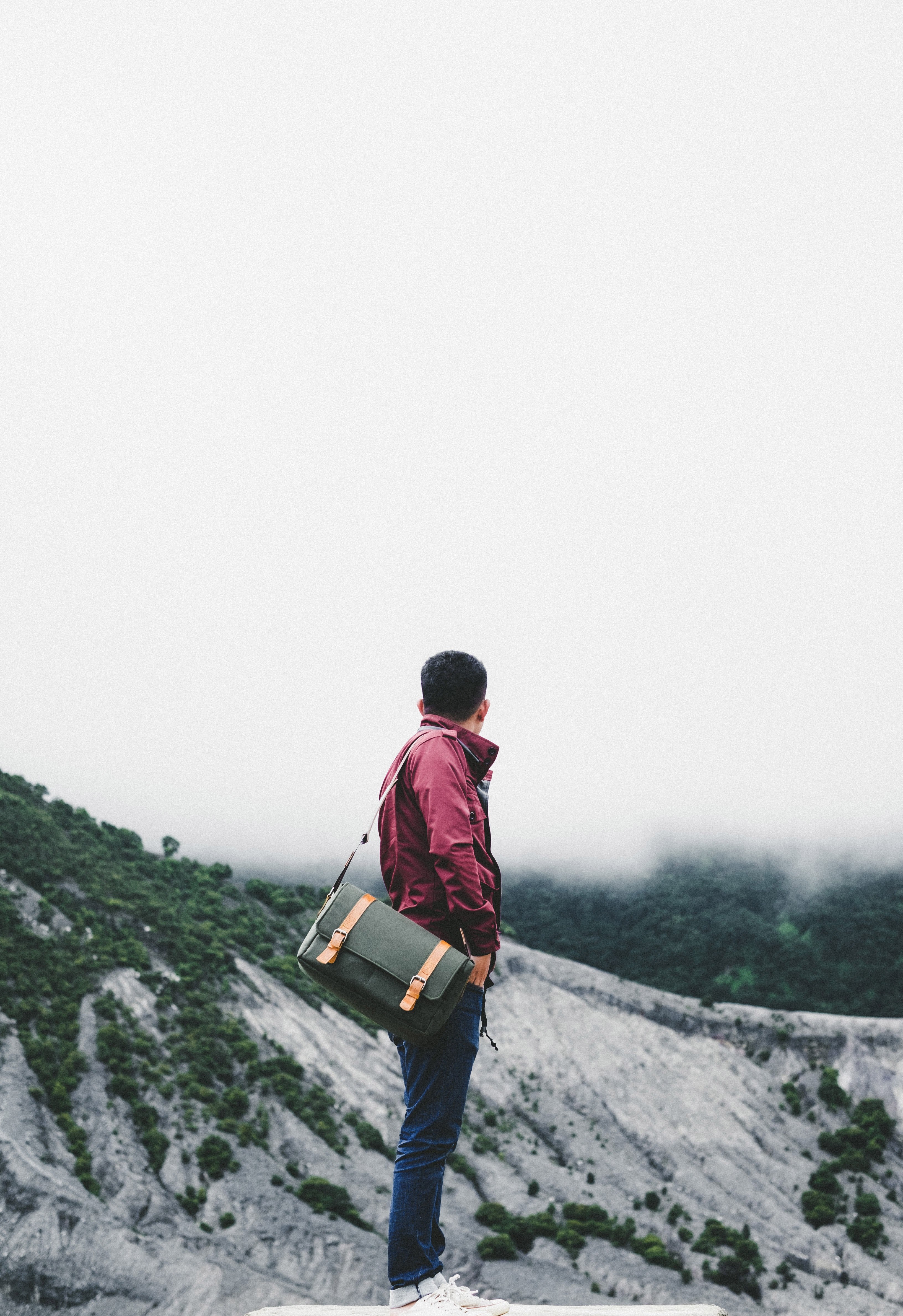 stylish man dressed in jeans and red jacket with leather satchel standing in gray mountain terrain, man red coat mountains 4k, Phone HD Wallpaper