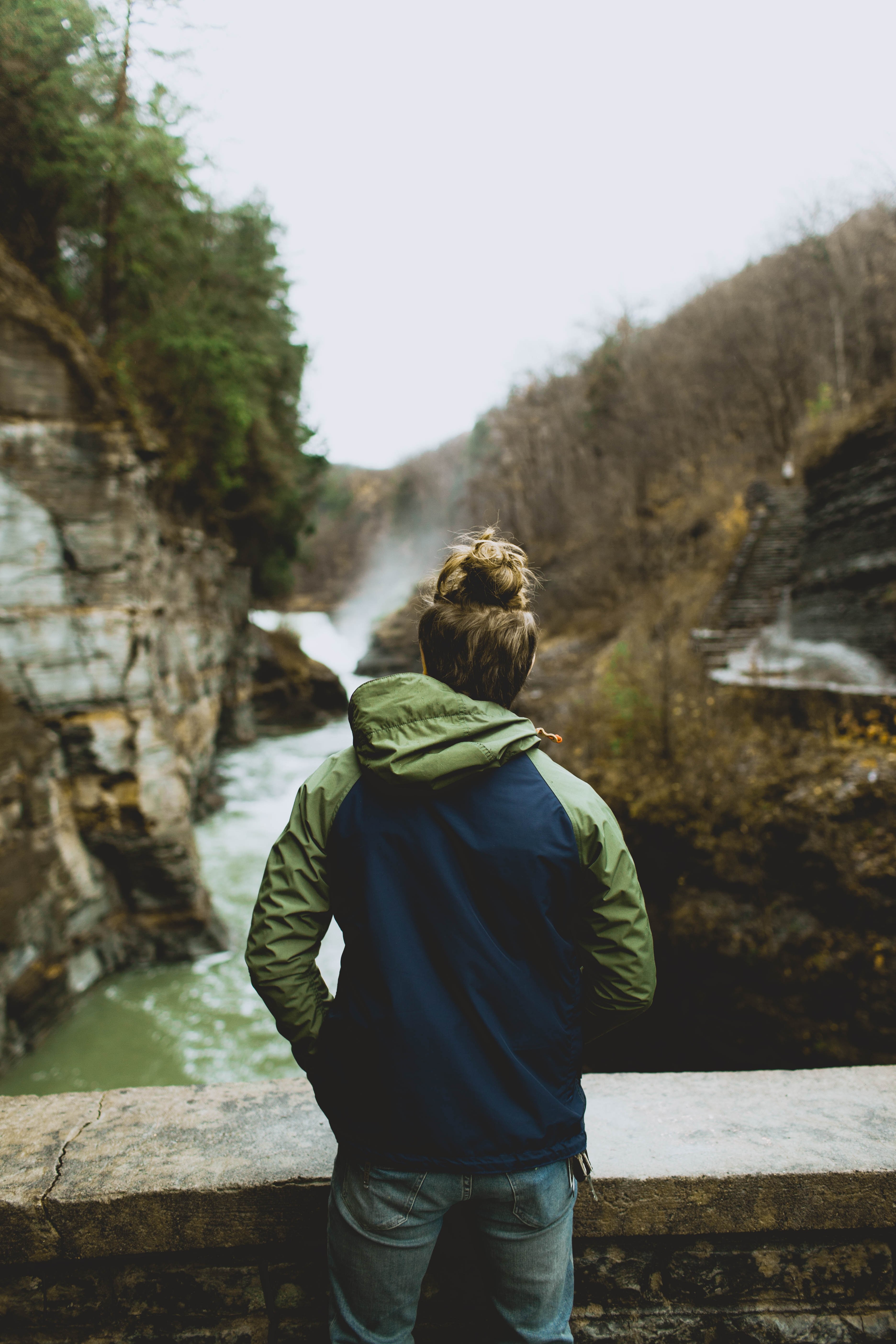 a woman in a jacket stands against a stone wall overlooking a river in the wilderness, gaze in wonder 4k, Phone HD Wallpaper