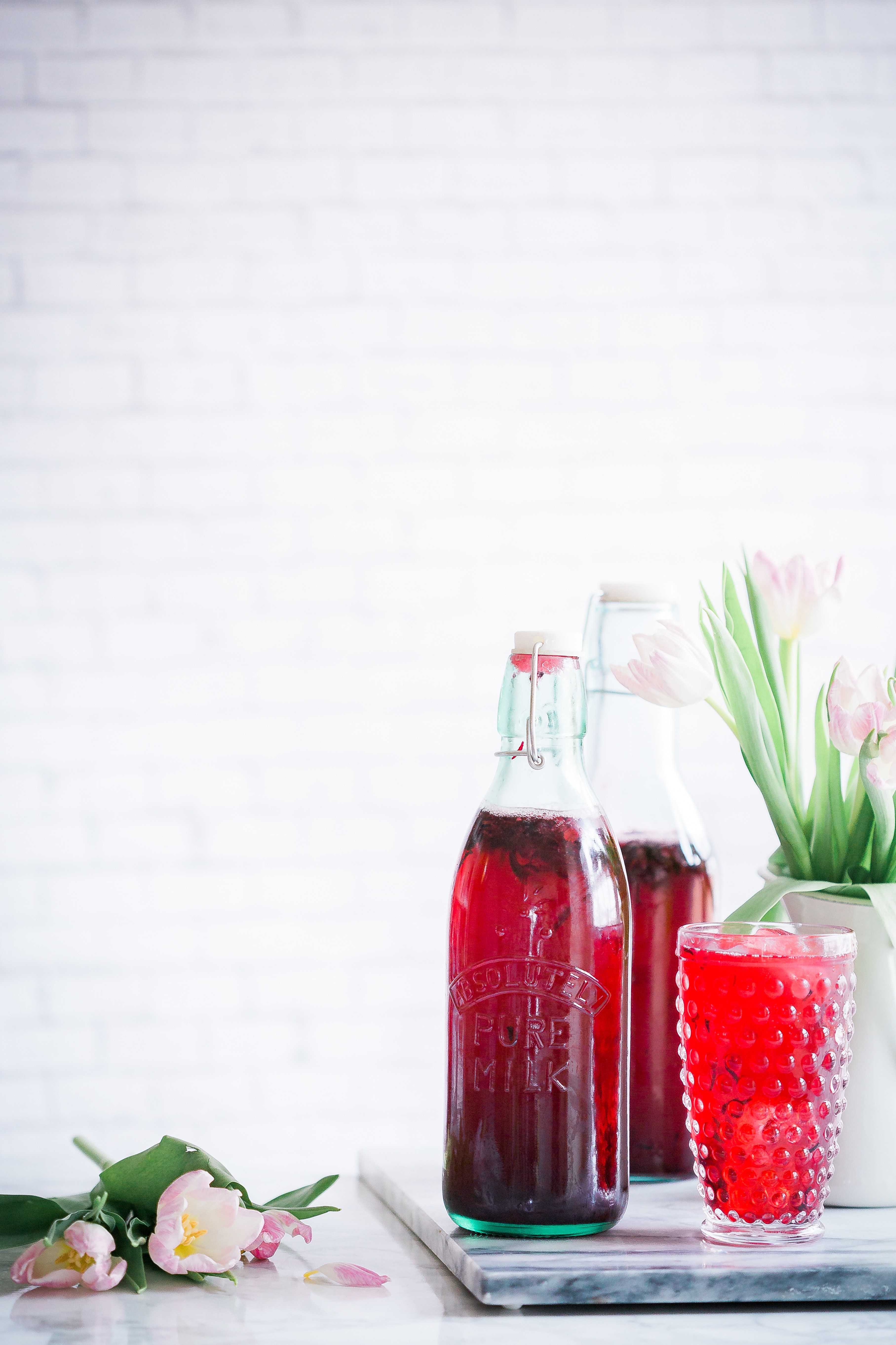 a red beverage in a glass and two milk bottles next to pink tulips on a table, sparkling hibiscus lemonade 4k, Phone HD Wallpaper