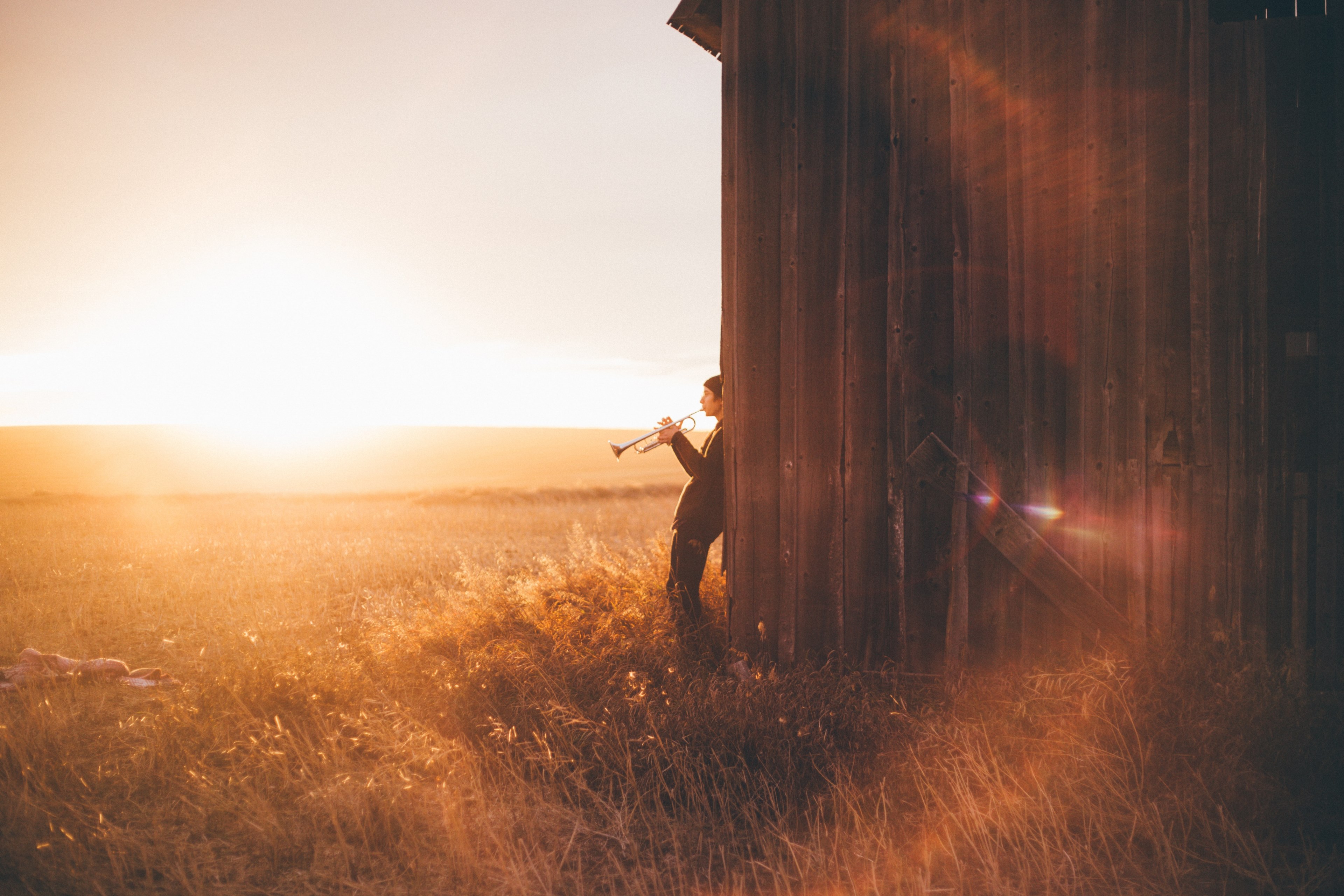 a man plays the trumpet leaning back against a barn wall and facing a golden field, trumpeter in a rural setting 4k, HD Wallpaper