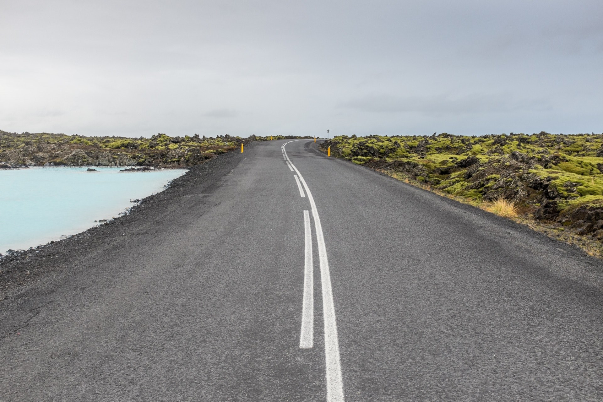a concrete road between light blue water and flat mossy rocks under a dull sky, driving through nature on a chilly morning 4k, HD Wallpaper