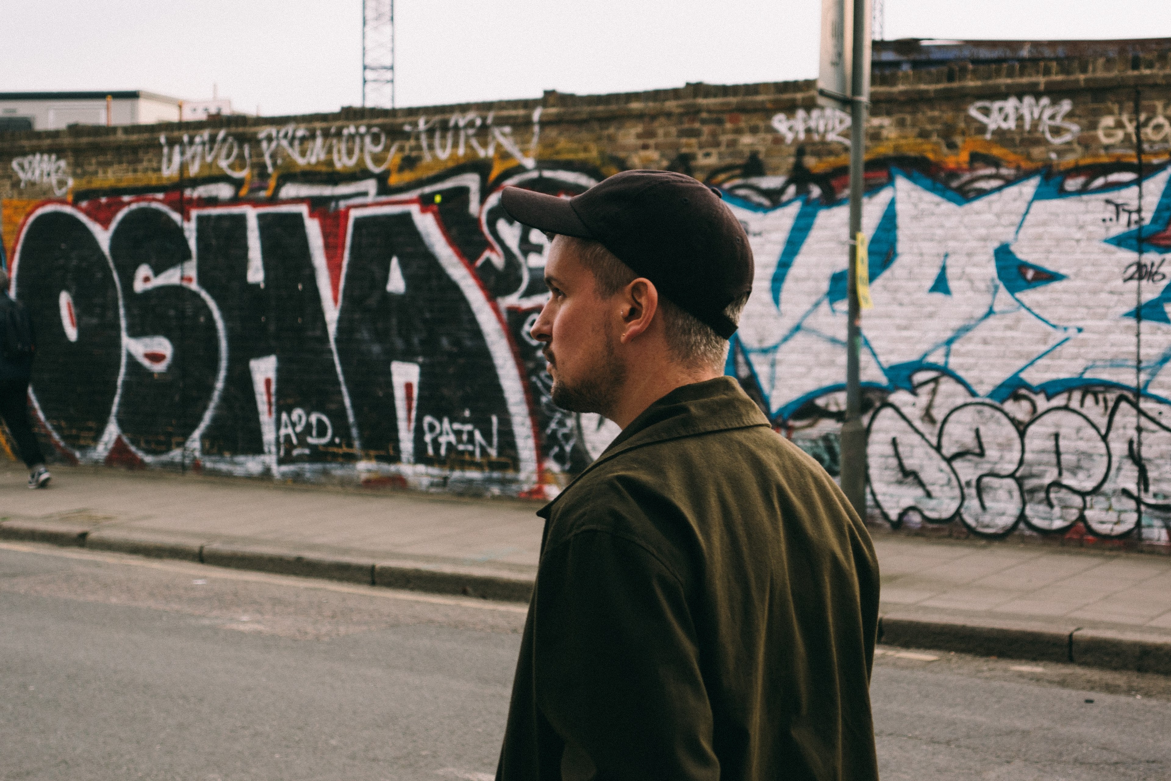 man wearing cap and jacket walks near hackney wick and passes graffiti, man walks in front of graffiti wall 4k, HD Wallpaper