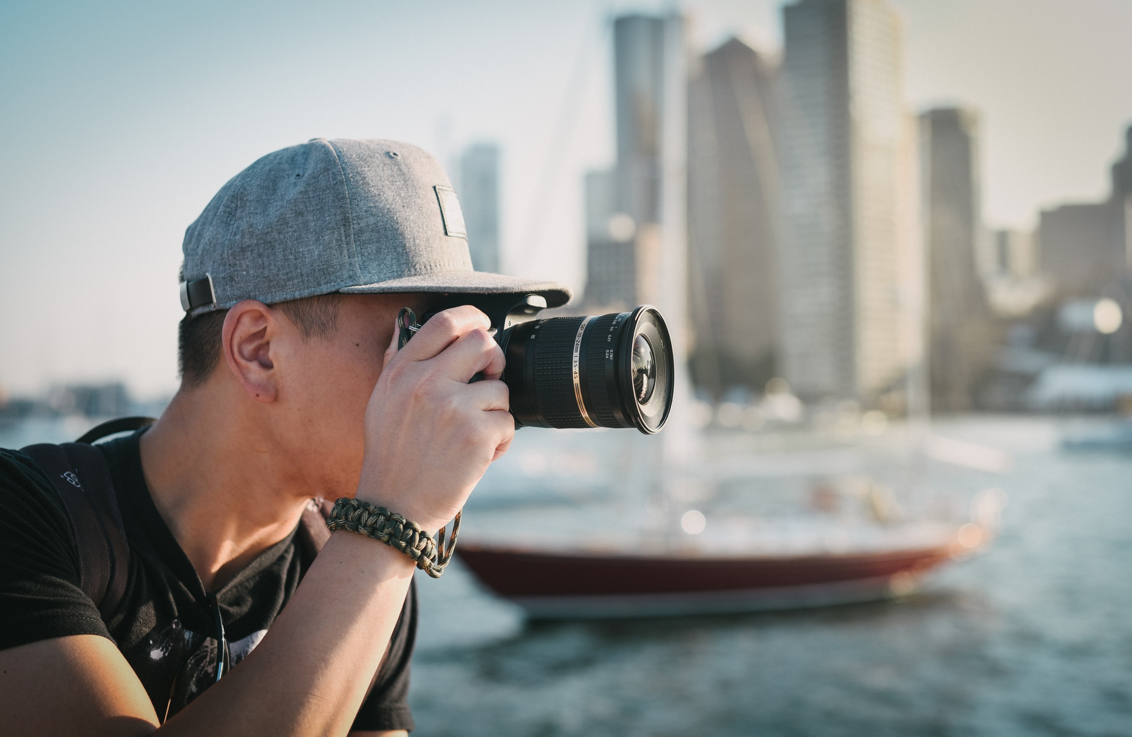a man wearing a cap and holding a camera with buildings and a boat beside him, man with camera 4k, HD Wallpaper