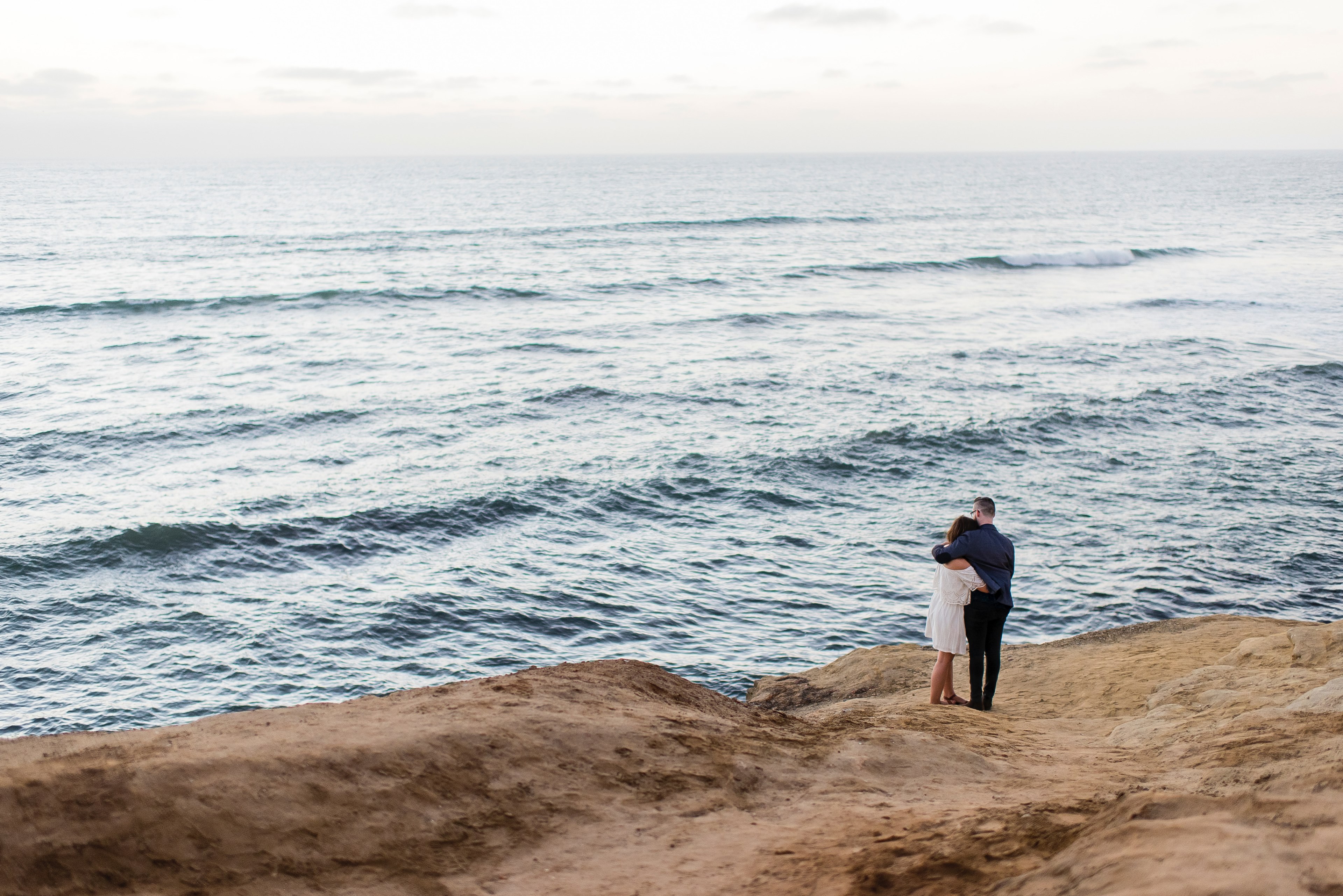 a couple stands on a rocky surface by the ocean their arms around each other, couple embrace sunset cliffs 4k, HD Wallpaper