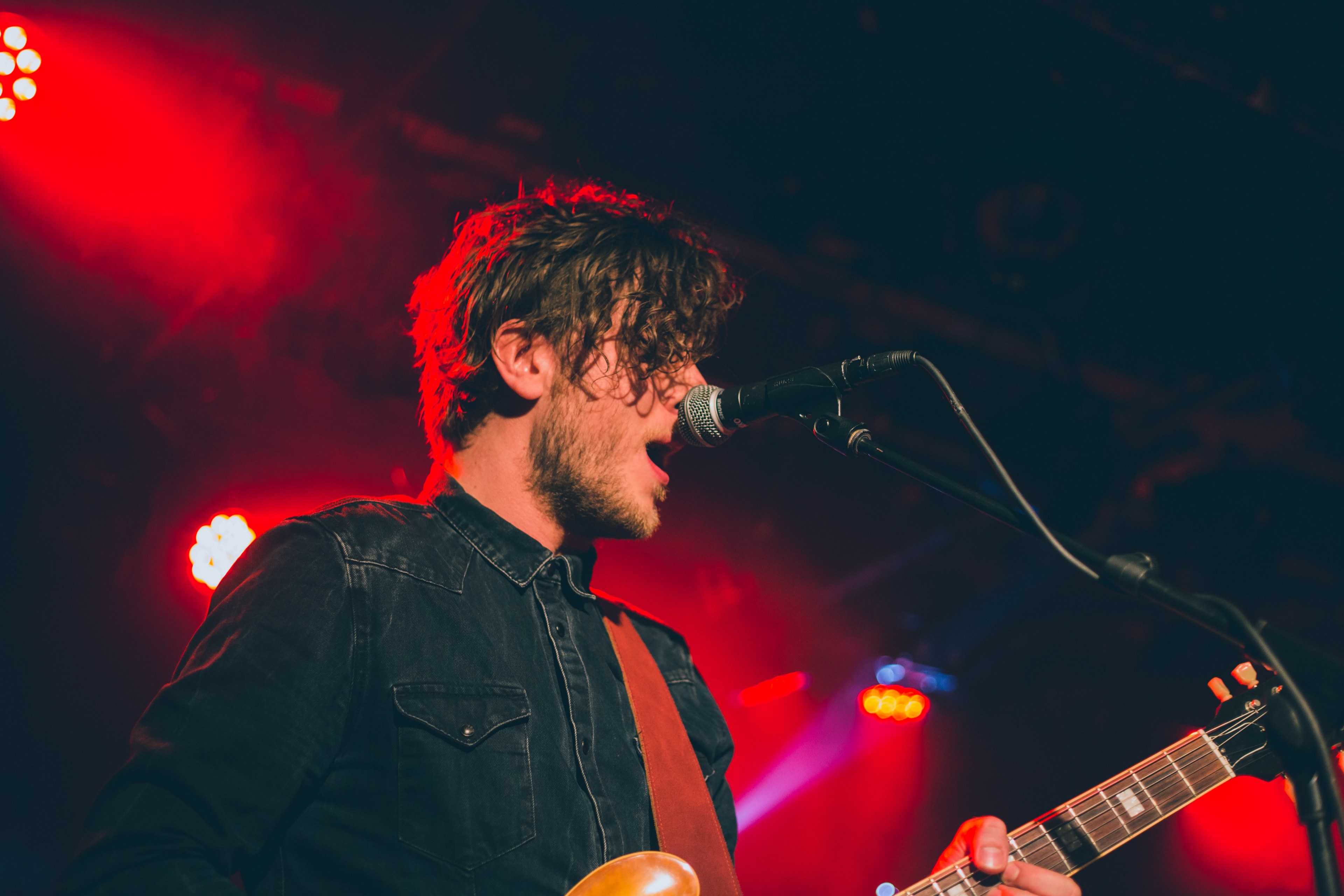 a bearded man with a guitar singing into a microphone with red lights behind his back, band member under red floodlights 4k, HD Wallpaper