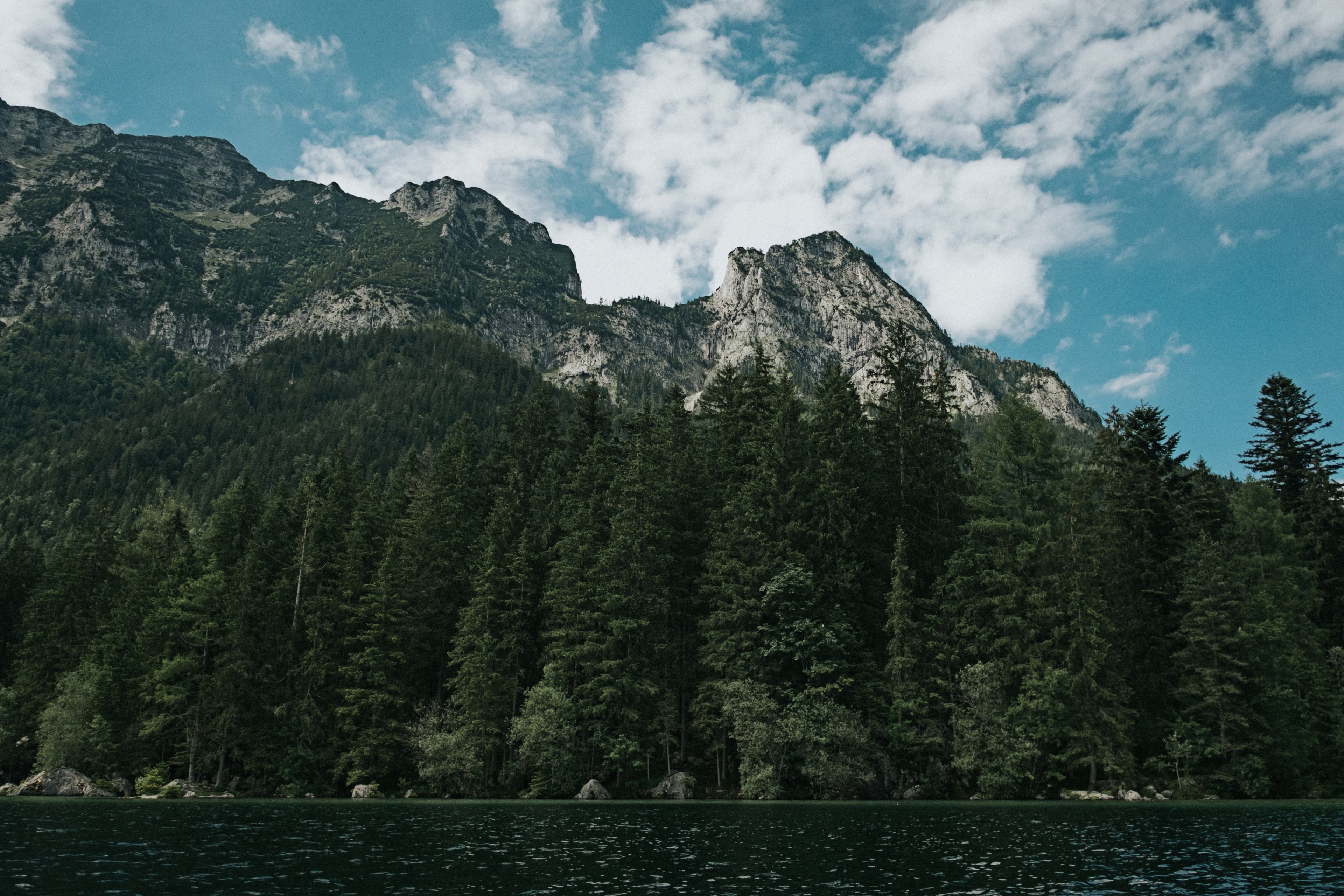 a forest surrounded lake at the foot of a craggy mountain range, mountaintops over the lake 4k, HD Wallpaper