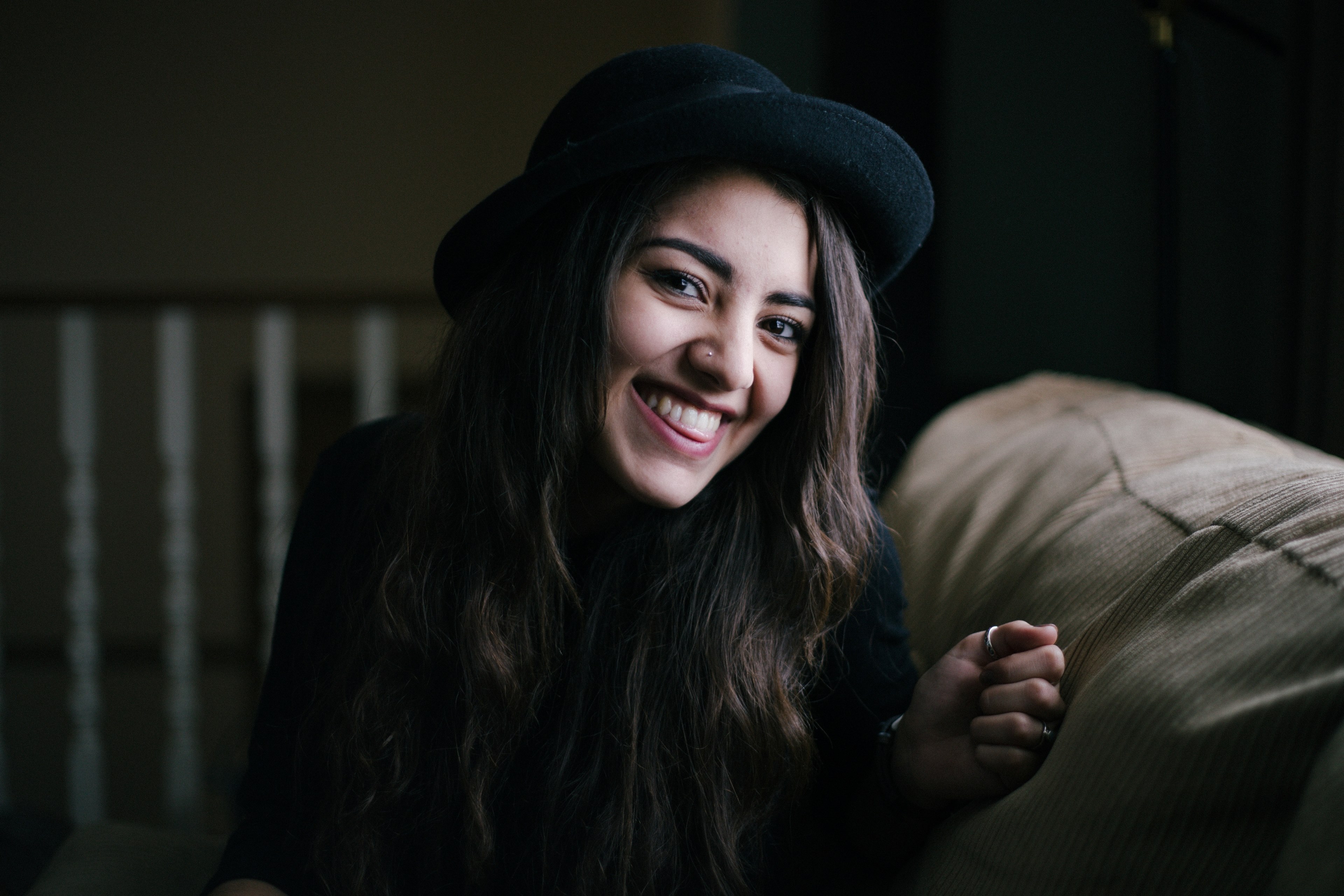 a woman with a black hat and nose piercing sits on a couch and smiles, woman in hat smiling on couch 4k, HD Wallpaper
