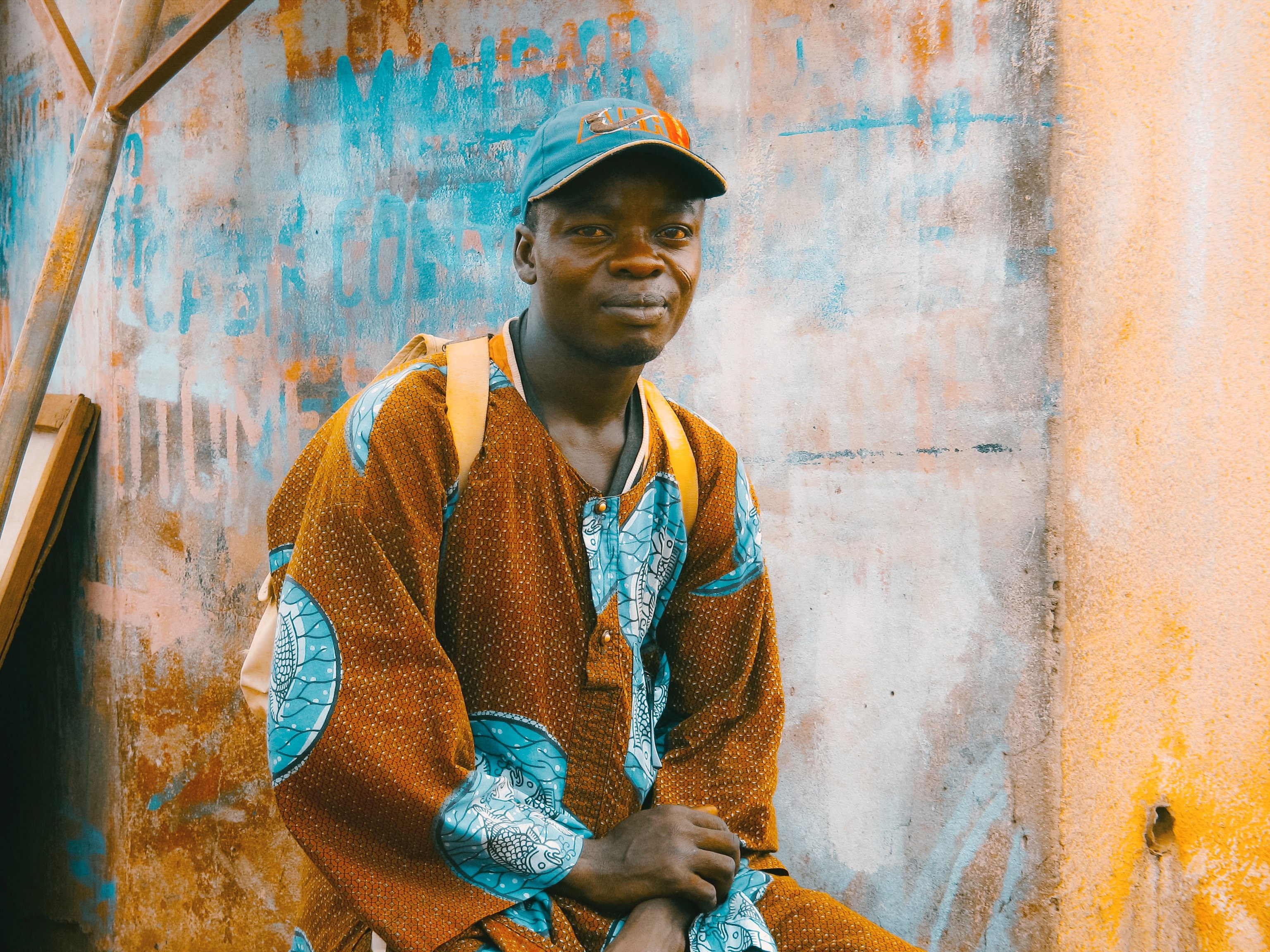 a man wearing a traditional orange and blue garment backpack and baseball hat near a graffitied wall, man in traditional outfit 4k, HD Wallpaper