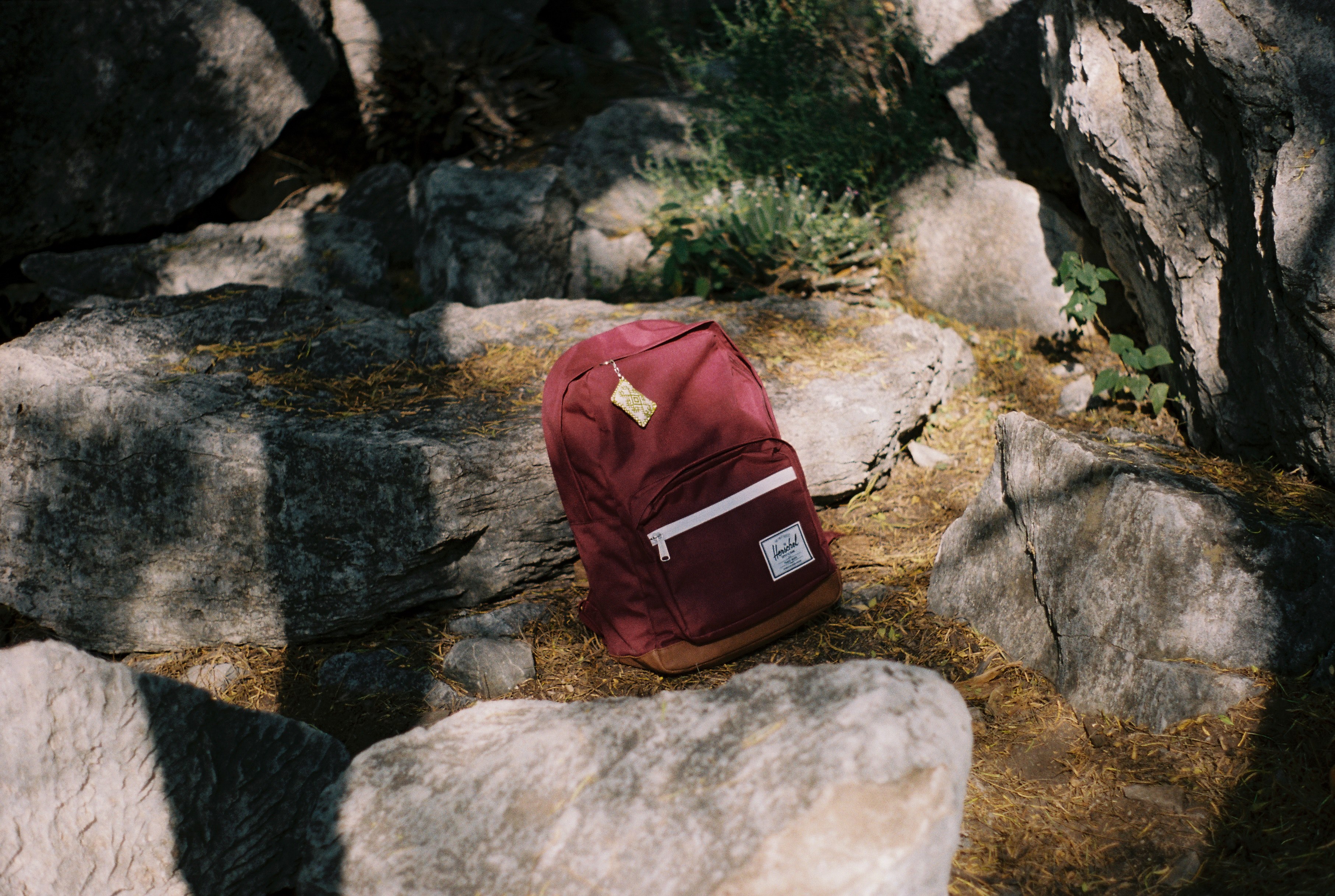 a burgundy herschel backpack with a coin wallet attached to the zip in the middle of a rocky outdoor area, herschel backpack by rocks 4k, HD Wallpaper