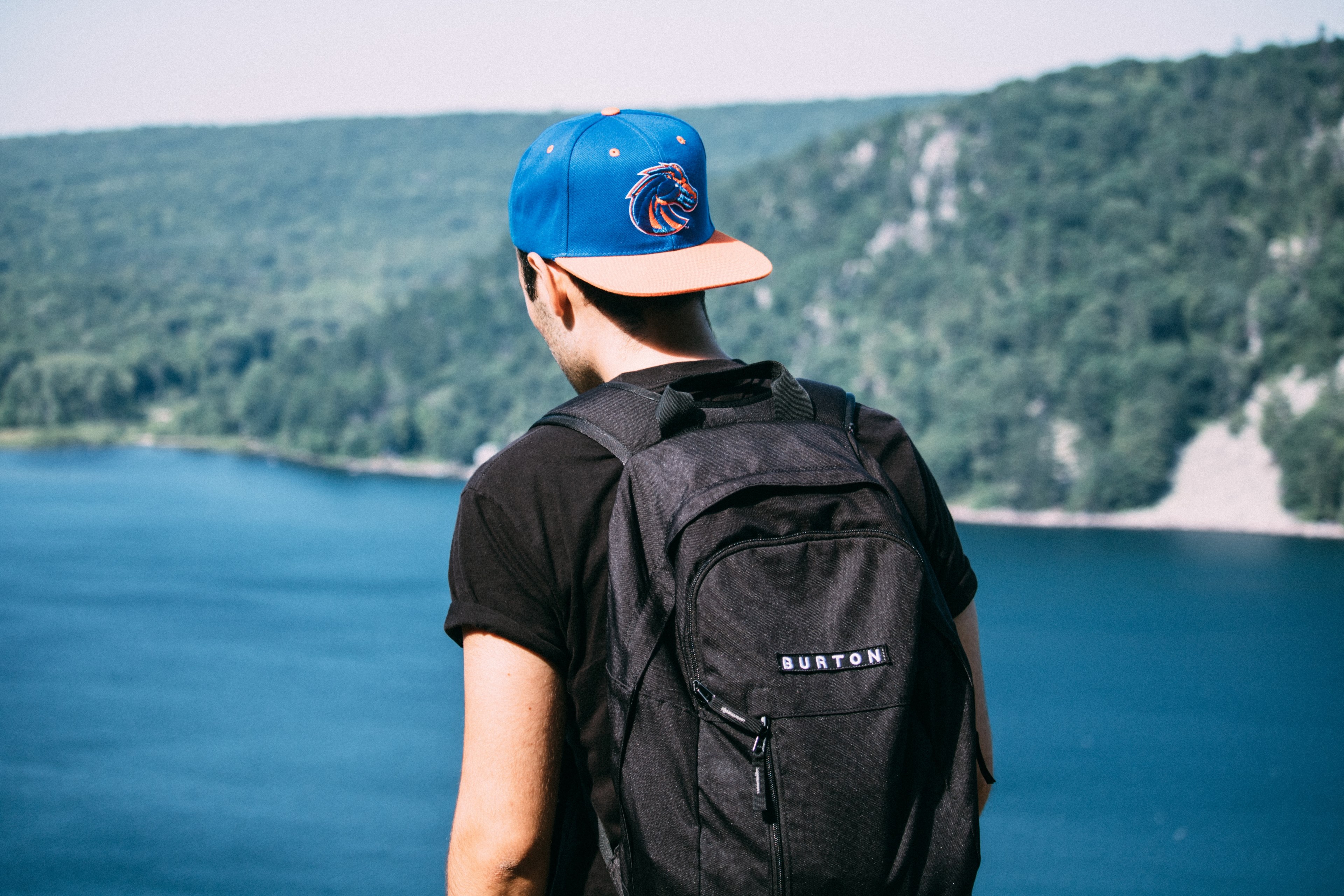 the back of a man wearing a backpack and cap backwards looking out in devils lake by forest of trees, man looking out to lake 4k, HD Wallpaper