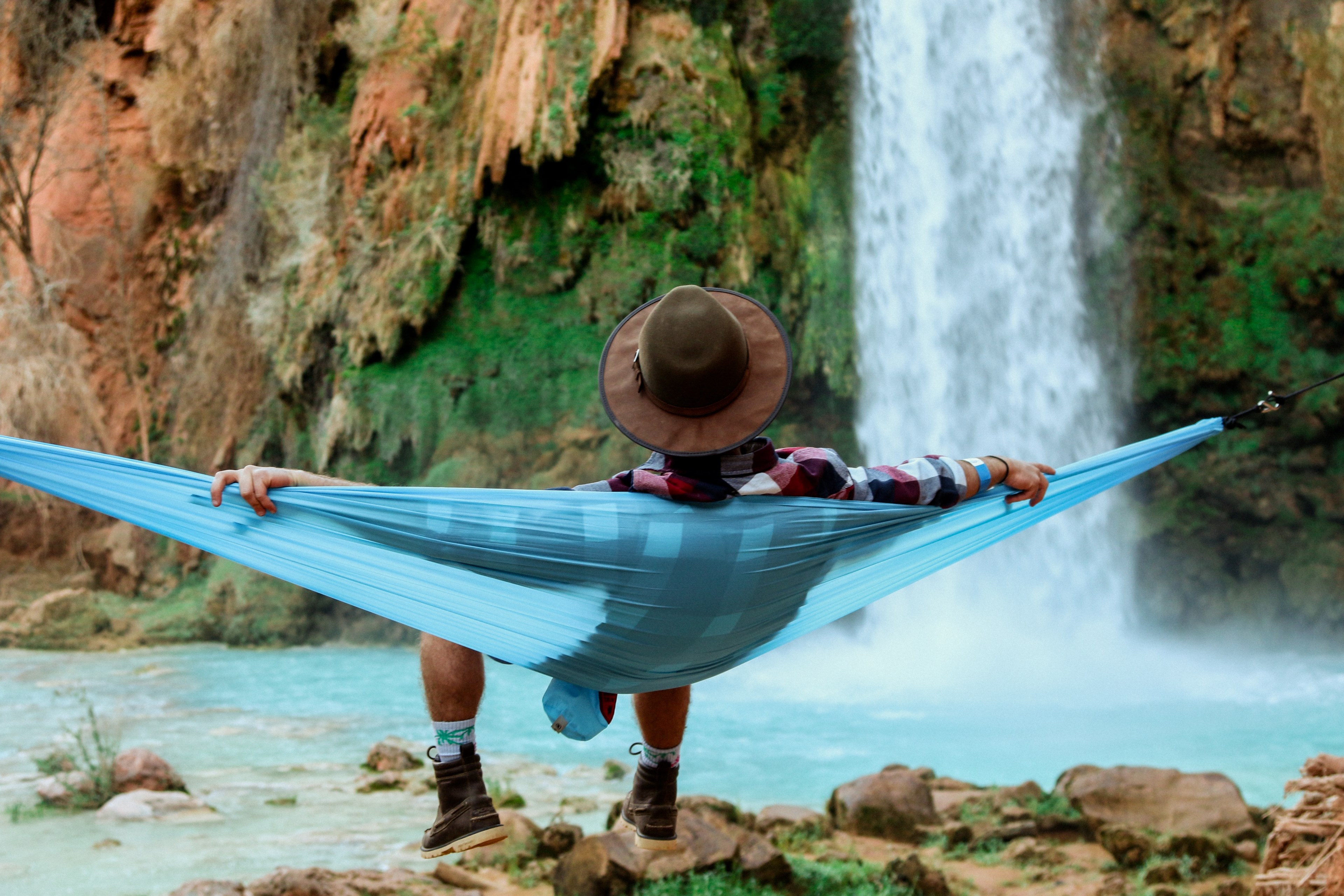 a man wearing a brown hat plaid shirt and hiking boots sitting on a blue hammock in supai falls, lost in the canyon 4k, HD Wallpaper