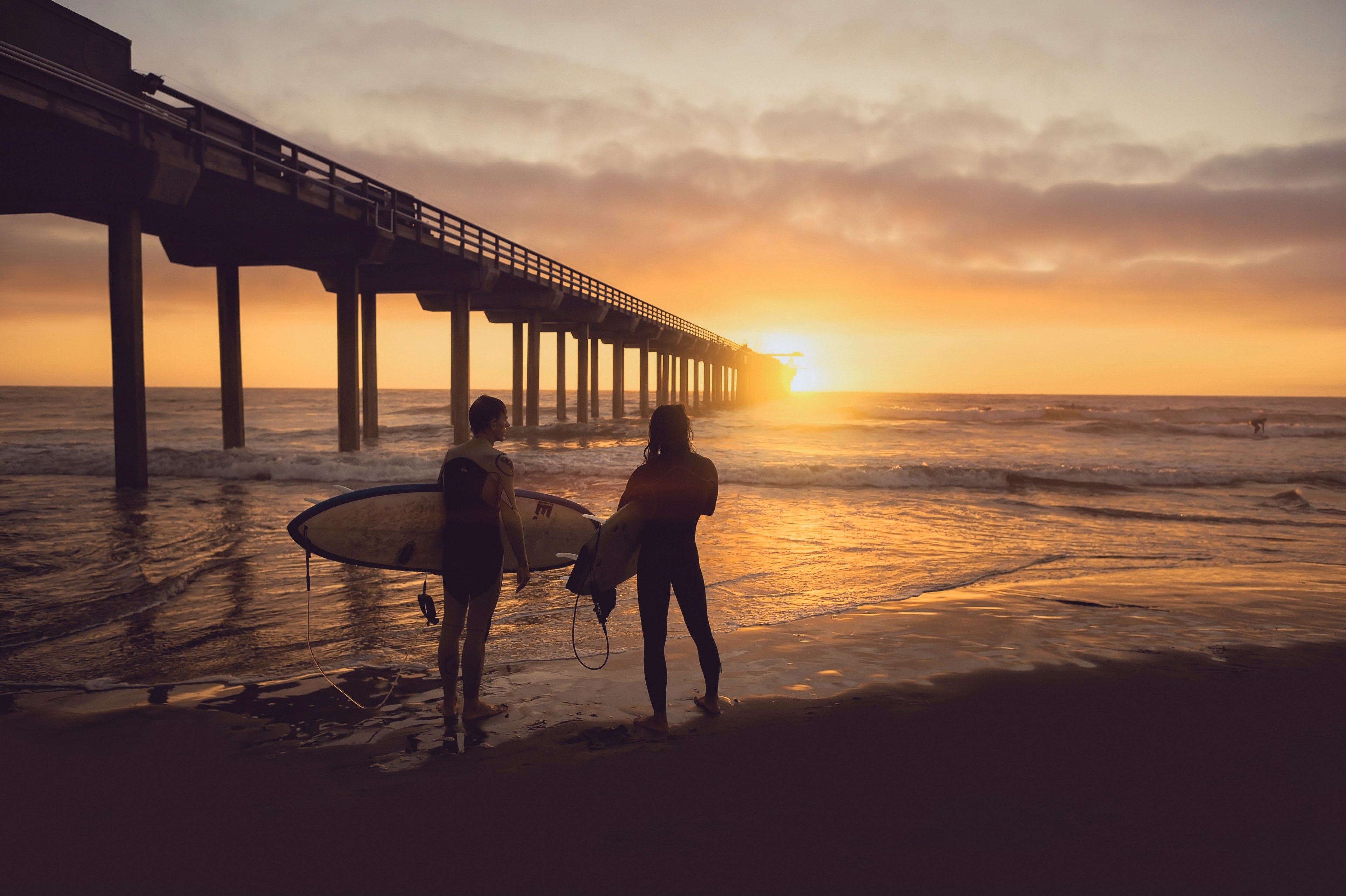 two surfers standing on the sand beach by the wooden pier pillars at sunset, surfing friends watching the sunset 4k, HD Wallpaper