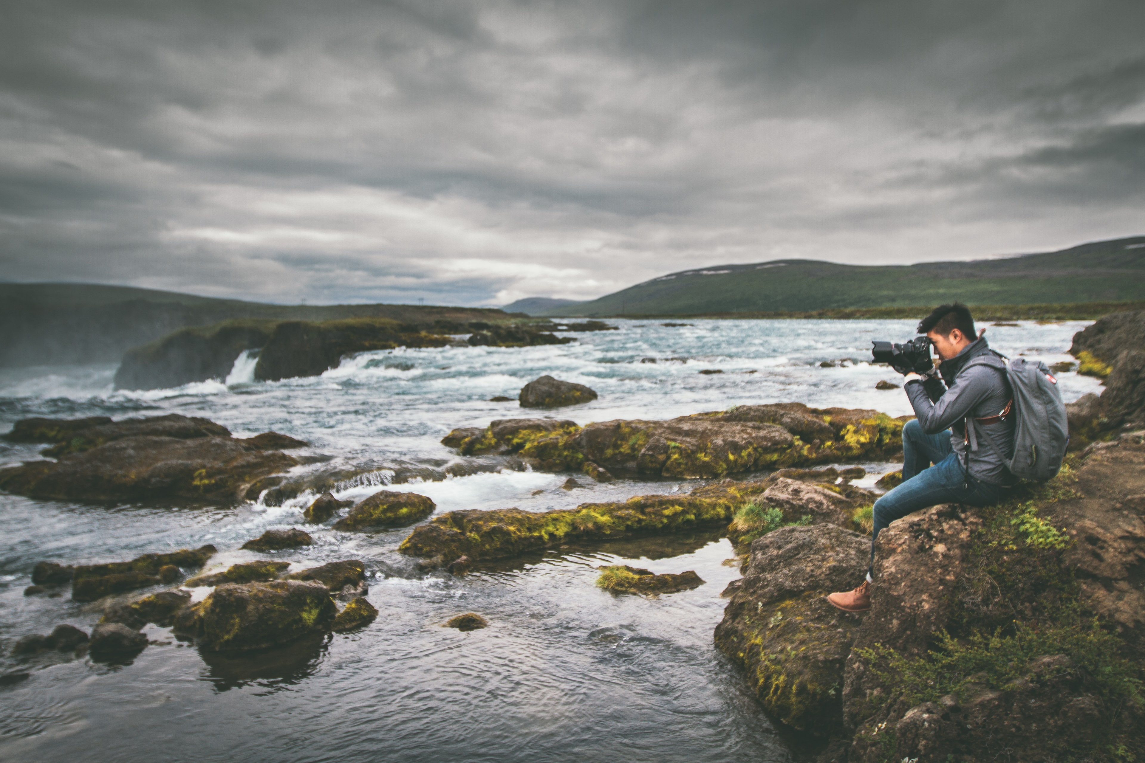 a man sits on a rock near the water to take a picture of the gafoss waterfall, man photographing waterfall 4k, HD Wallpaper
