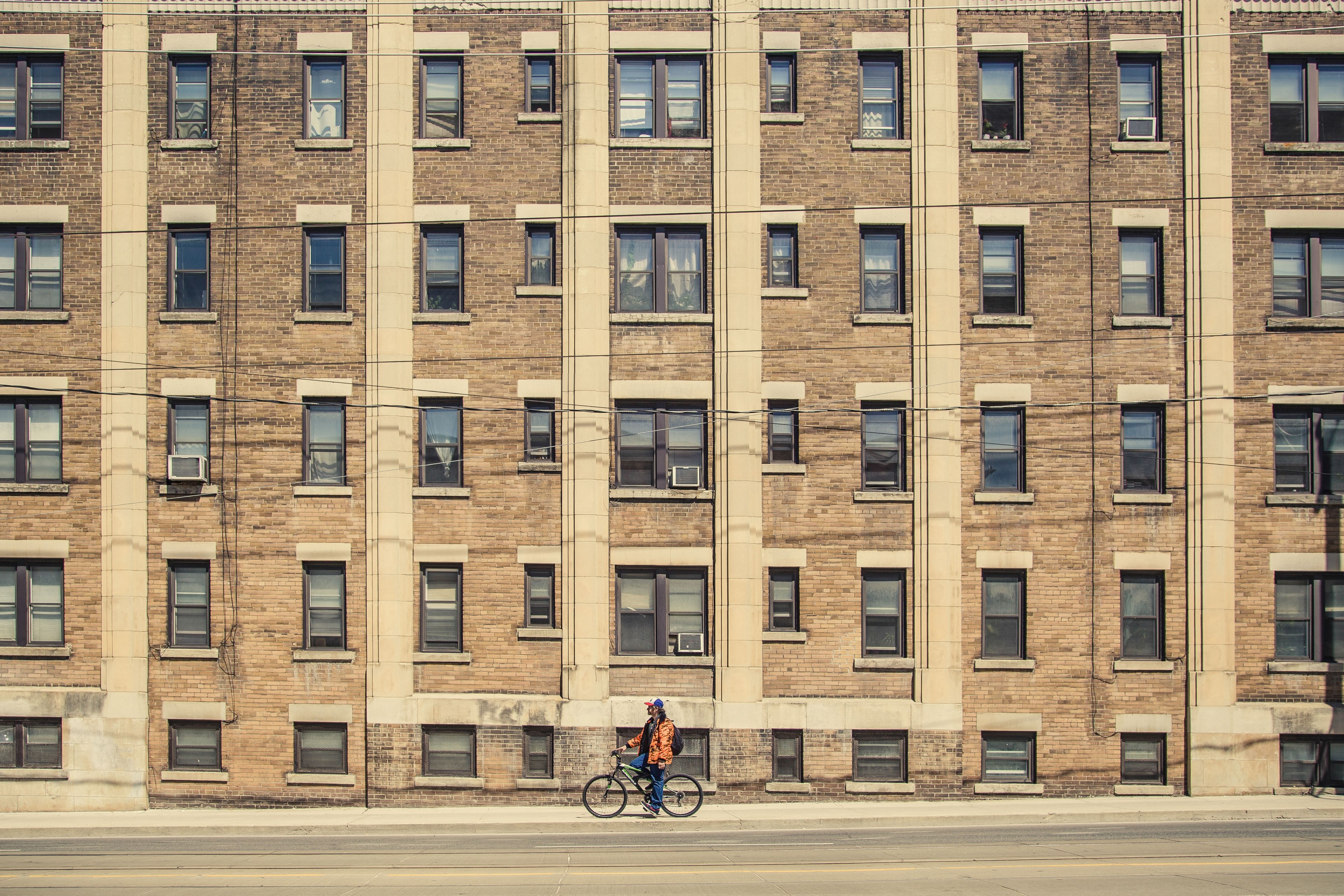 man in orange coat resting on bike in front of brown urban apartment building with windows, man on bike urban apartment 4k, HD Wallpaper