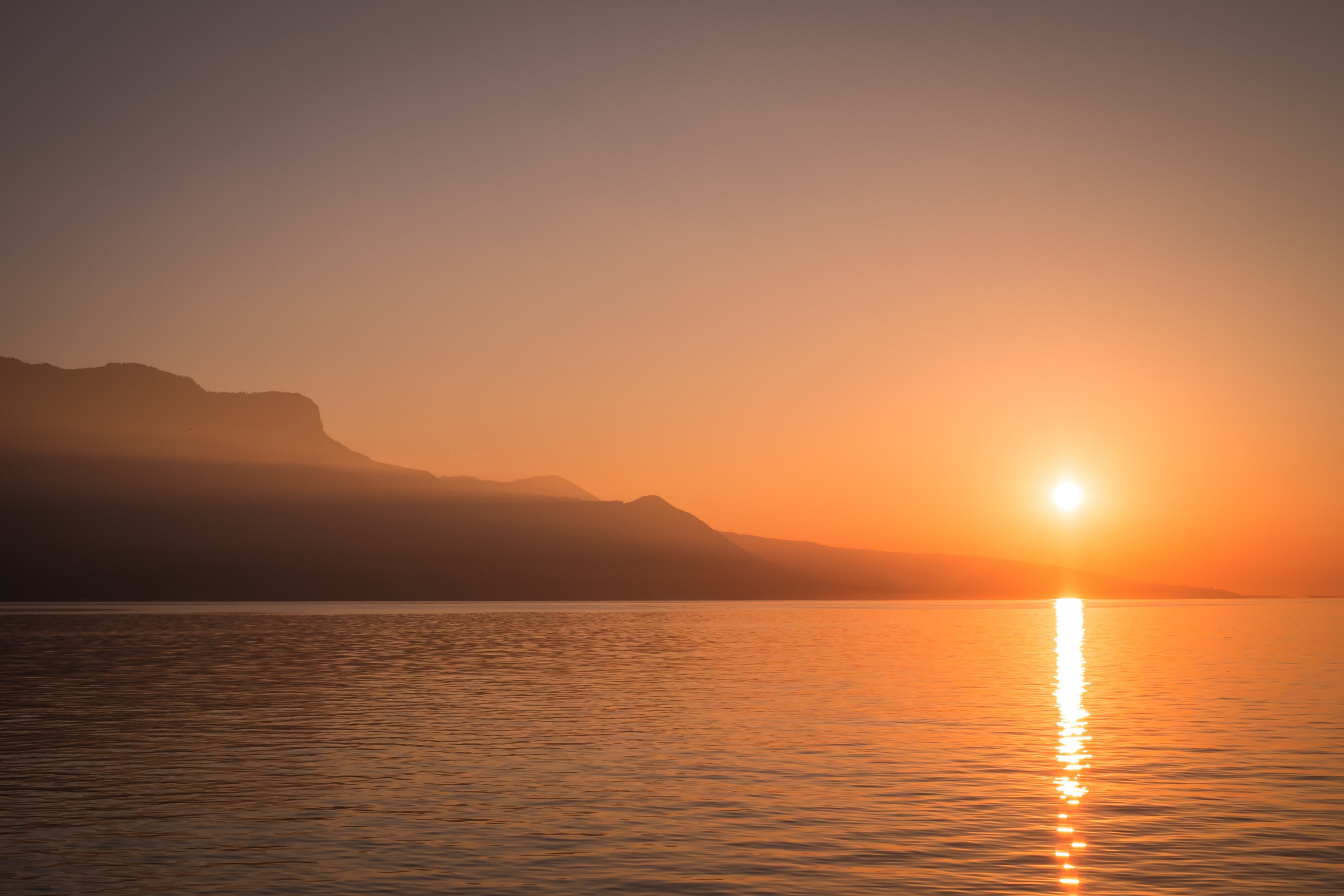 a sunset over a lake next to a silhouette of a steep rock face, sunset by a lake rock face 4k, HD Wallpaper