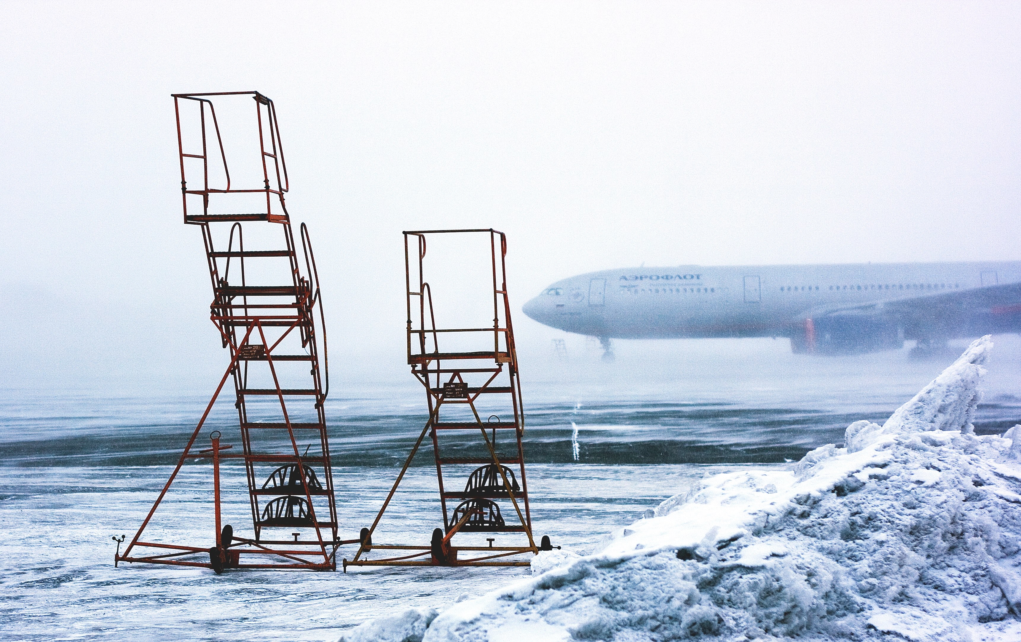 two sets of airstairs sit next to each other on a frozen tarmac a grounded plane is visible in the background, airstairs on snowy tarmac 4k, HD Wallpaper