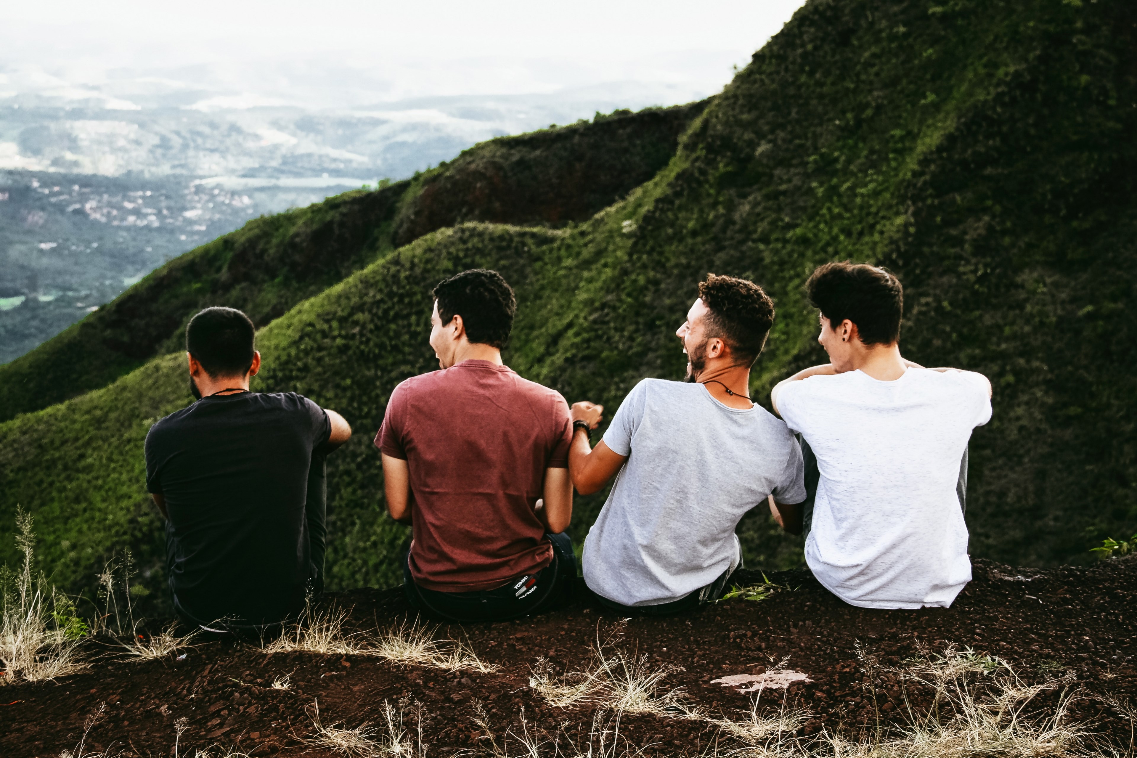 a group of four laughing men sits on an edge overlooking a green valley, men laughing by green valley 4k, HD Wallpaper