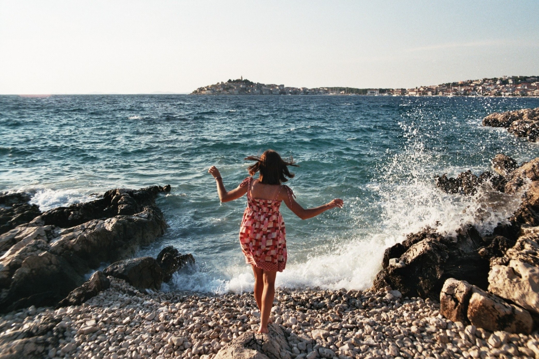 a woman in a dress standing on a rock on the coast with waves crashing against the nearby rocks, sea breeze and splashing waves 4k, HD Wallpaper
