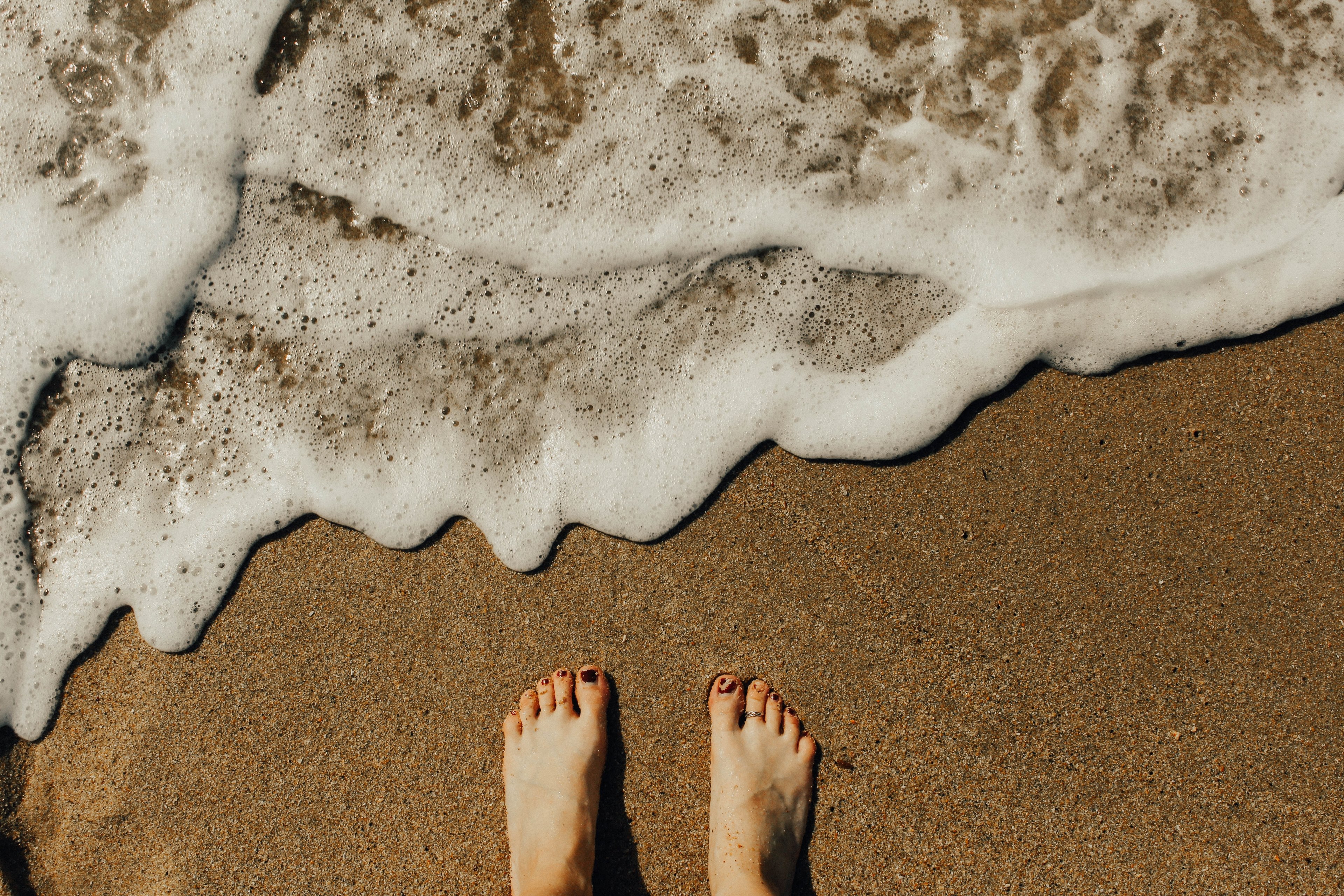 an overhead shot of a persons feet in wet coarse sand next to small foaming waves, feet in the wet sand 4k, HD Wallpaper