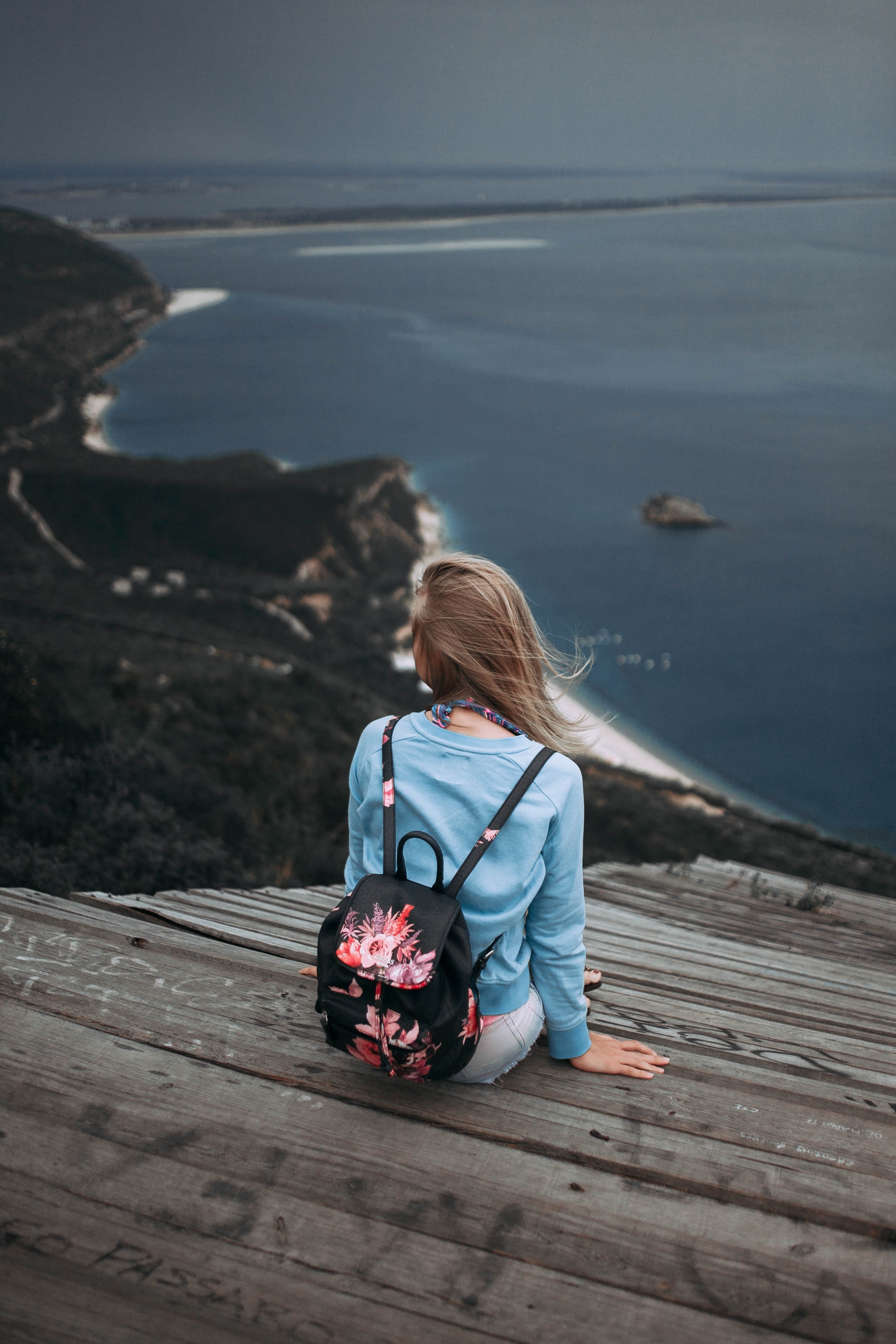 a woman with a backpack sitting on a wood surface while staring out into the ocean, _windy day 4k, Phone HD Wallpaper