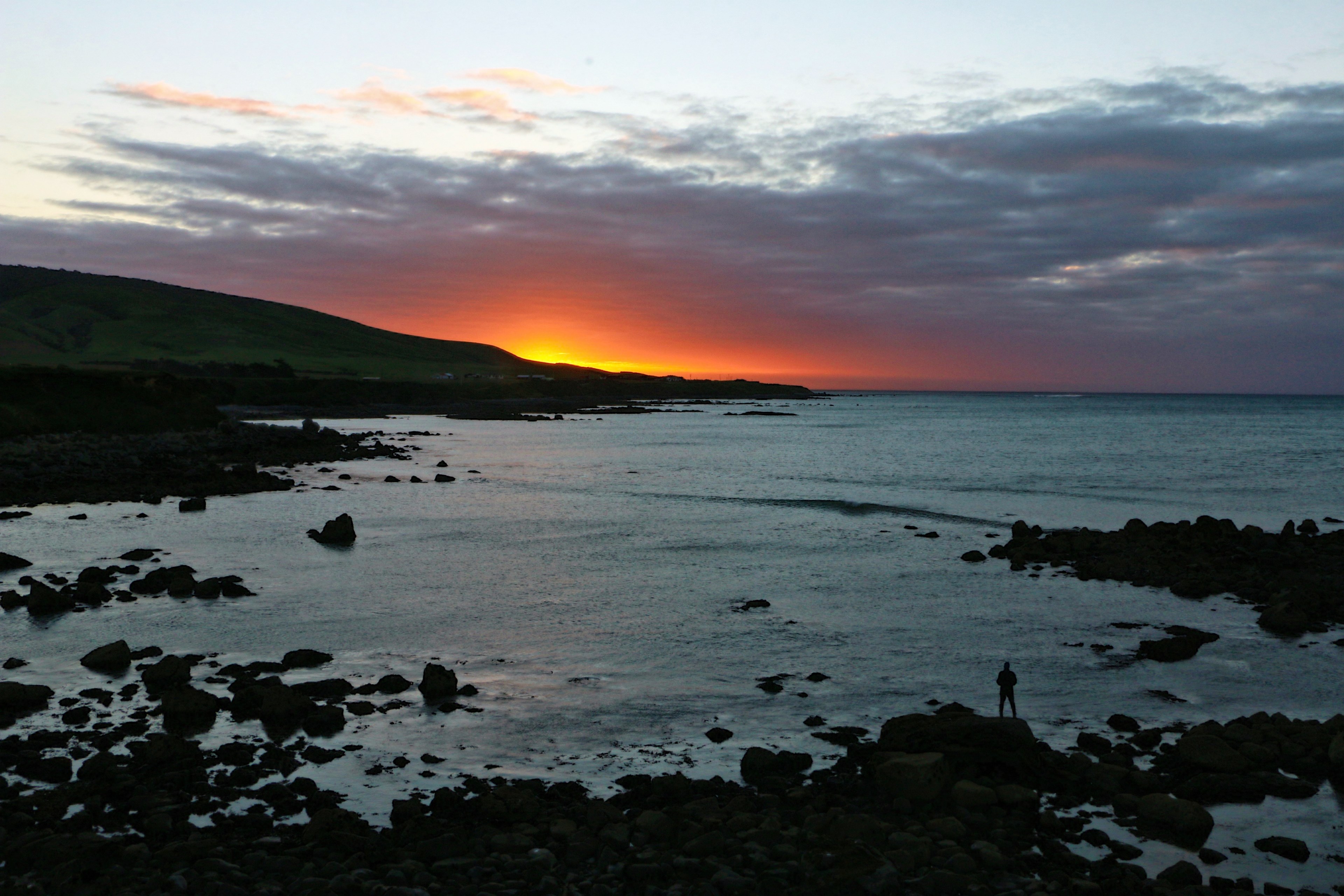 distant shot of a person standing on a rocky beach as clouds gather sun sets behind a far mountain, sunset at monkey island 4k, HD Wallpaper