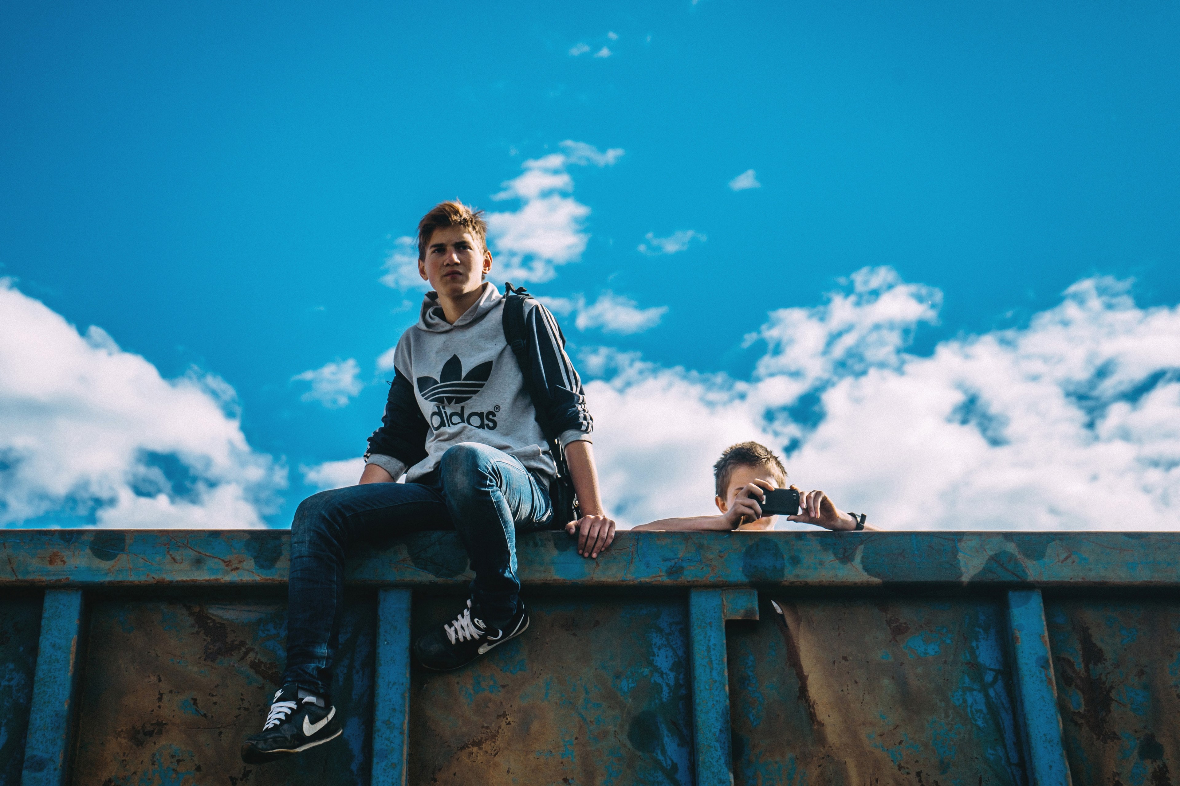two men on a rusted wall against a blue sky one taking photos with an iphone, men on rusted wall 4k, HD Wallpaper