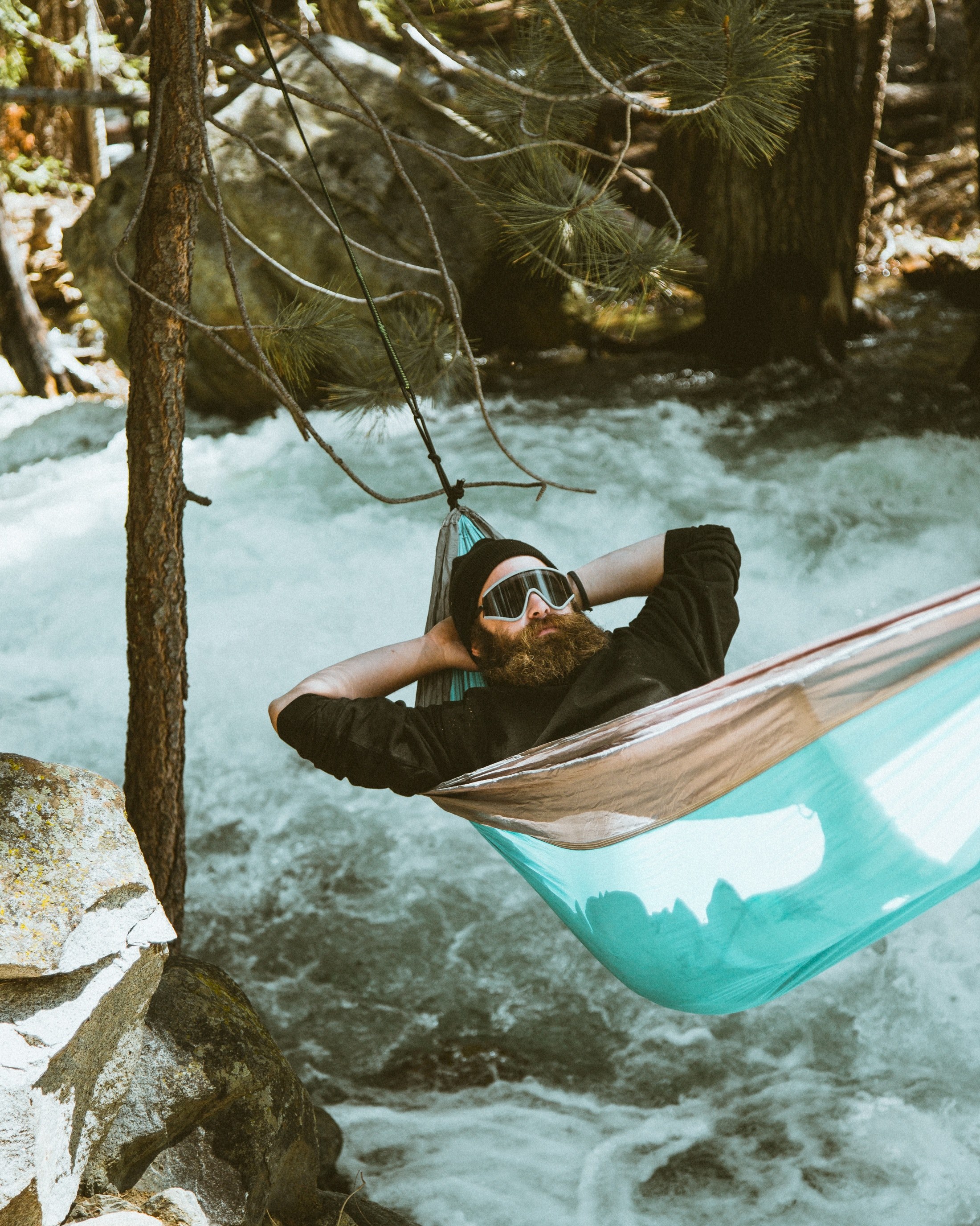 a man with a beard wearing goggles and a hat laying on a blue hammock over a river in the forest, hanging out over the river 4k, Phone HD Wallpaper