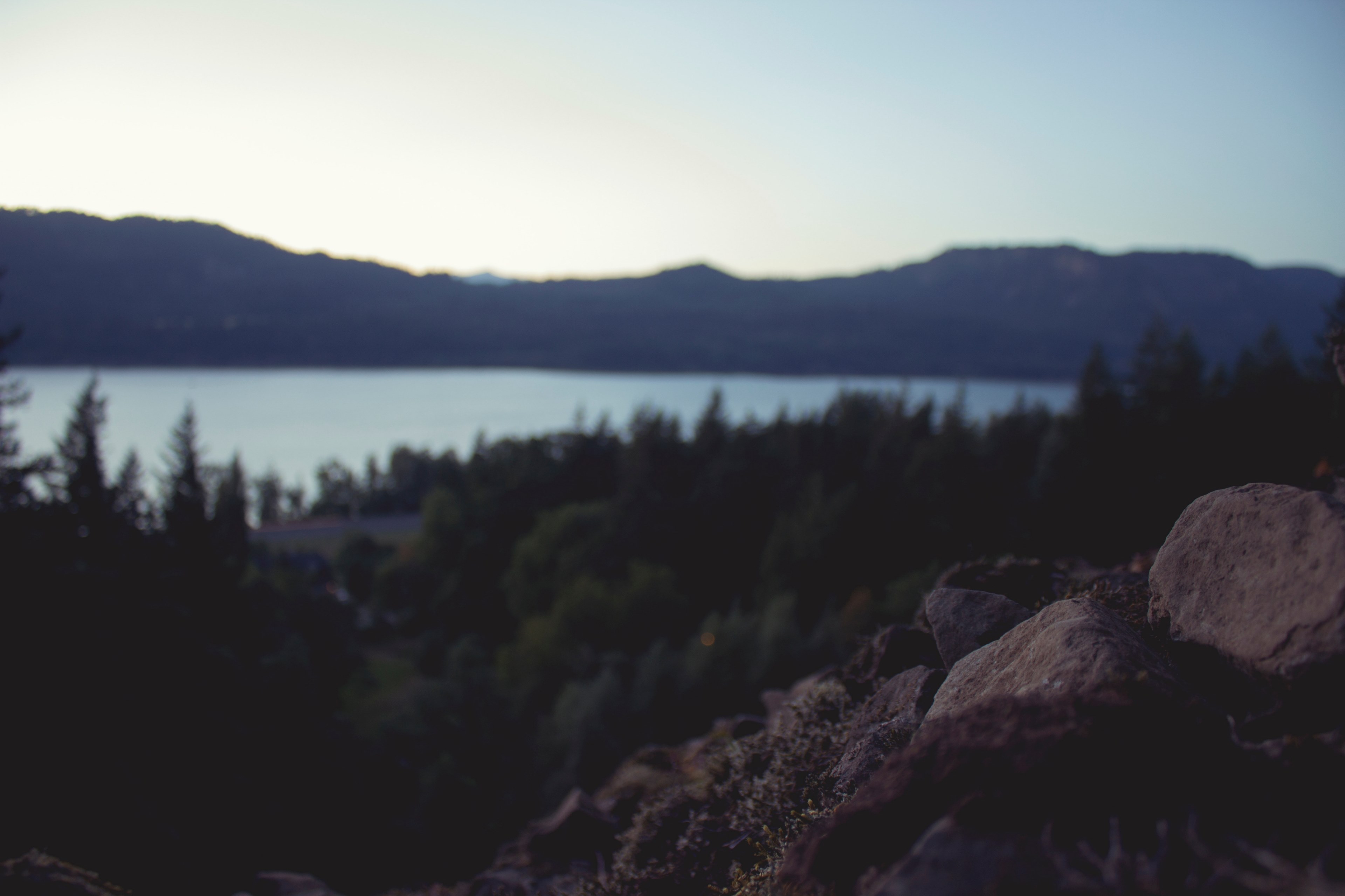 a lake encircled by a forest partially obscured by brown rocks in the foreground, lake in the distance 4k, HD Wallpaper